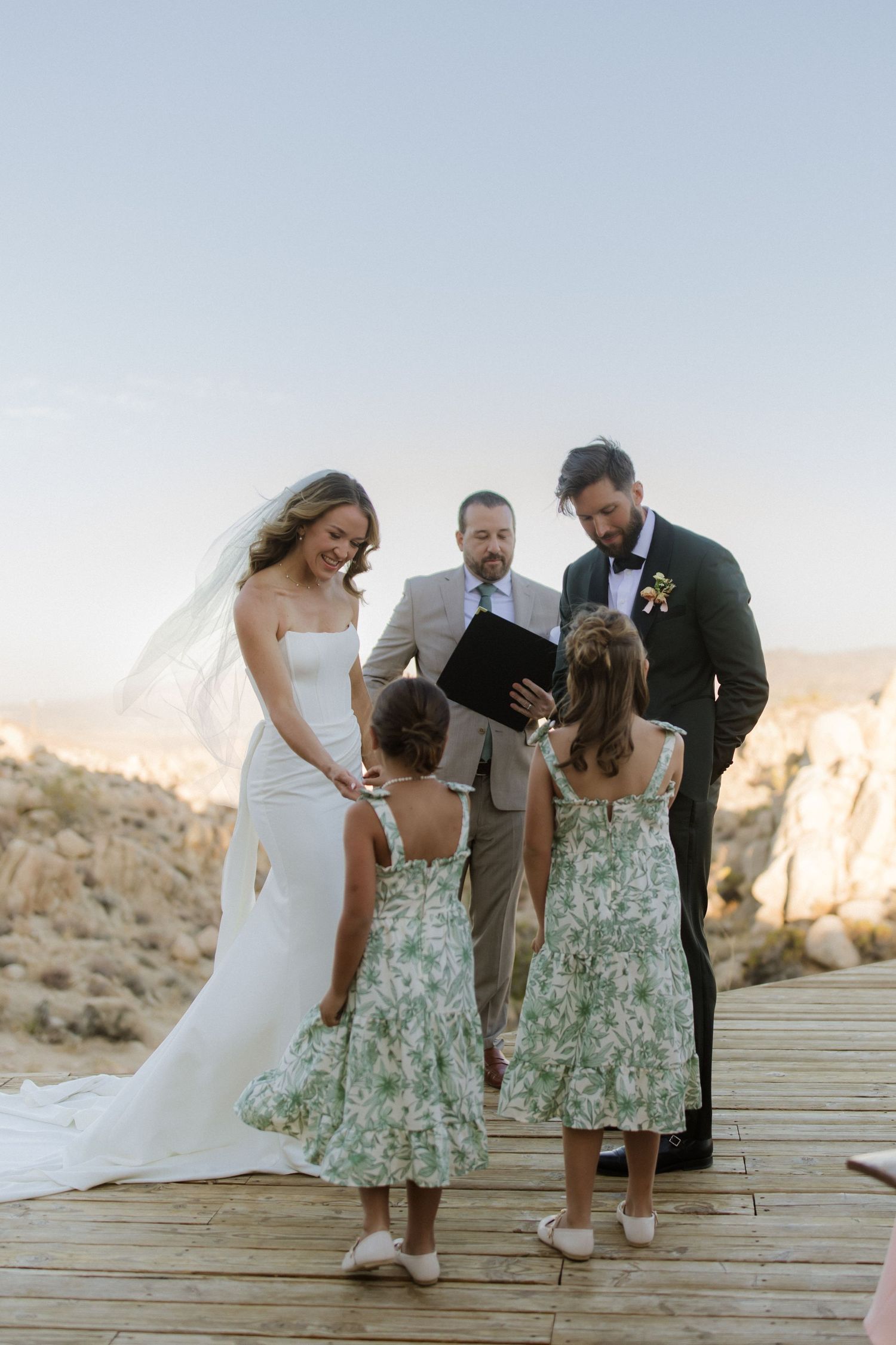 A wedding group stands on wooden decking in the desert at sunset, sharing heartfelt moments during a ceremony.