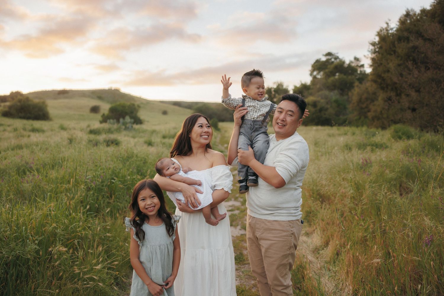 Family of four poses in a green meadow at sunset, parents holding two young children, smiling joyfully.