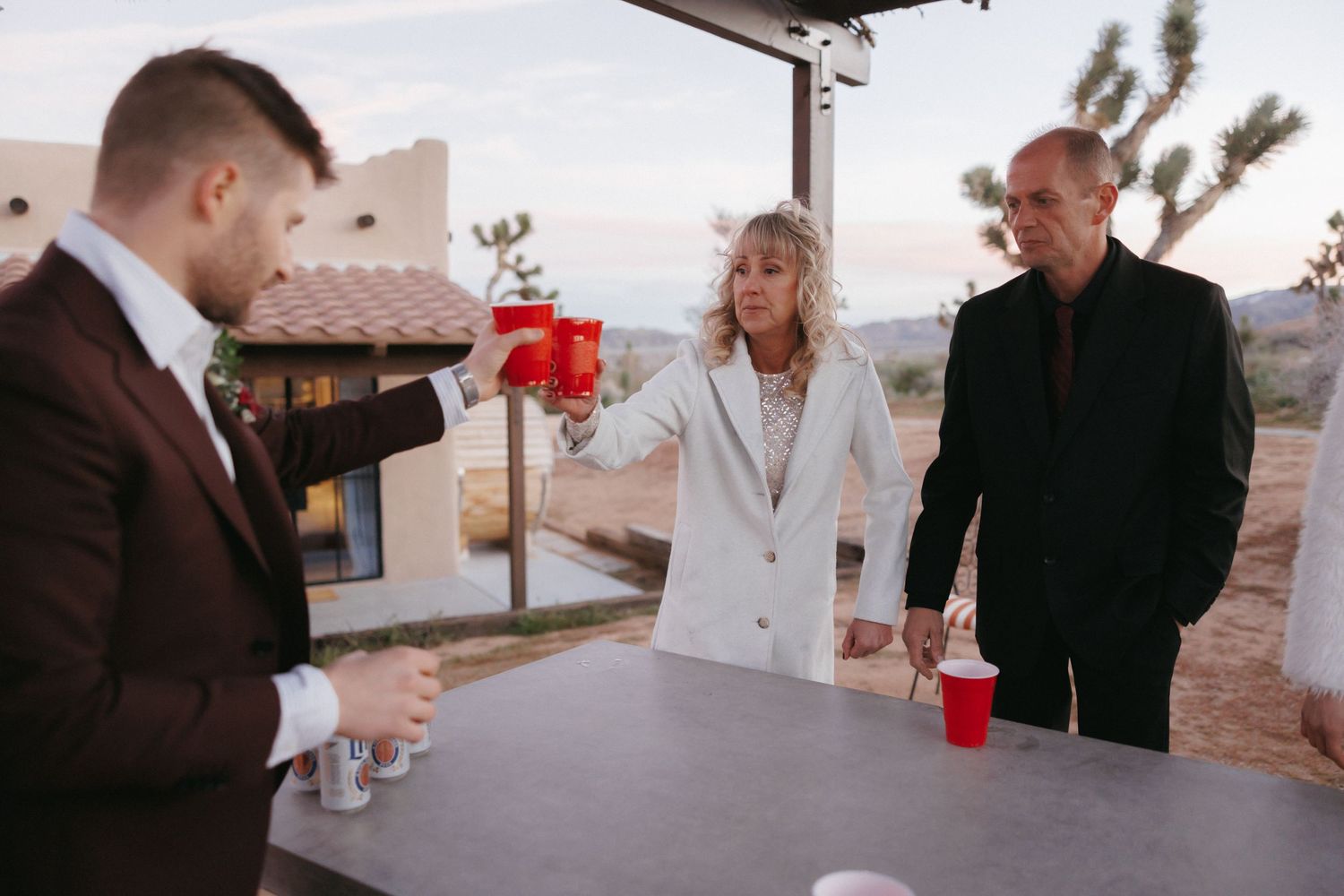 Groom in burgundy suit toasts with guests at outdoor desert wedding reception bar at dusk.