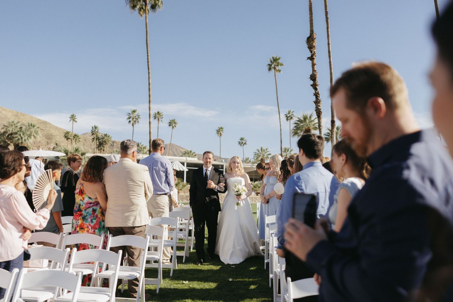Bride and groom walking down aisle at outdoor wedding ceremony surrounded by palm trees and guests.