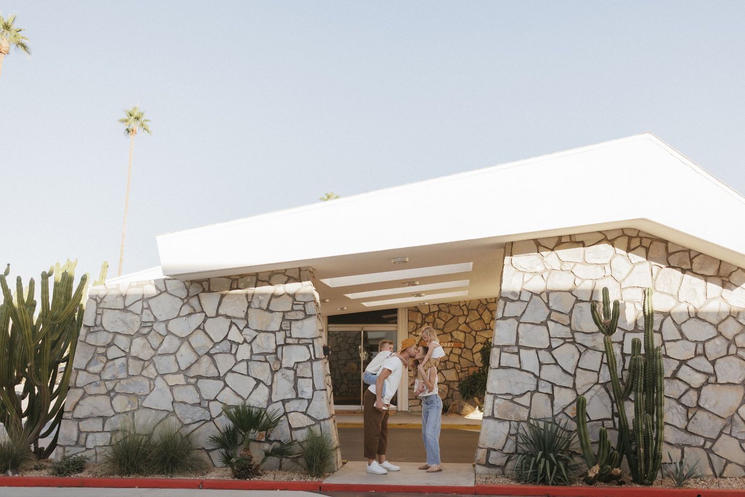 Two people standing outside a mid-century modern stone building with cacti and palm trees in Palm Springs.