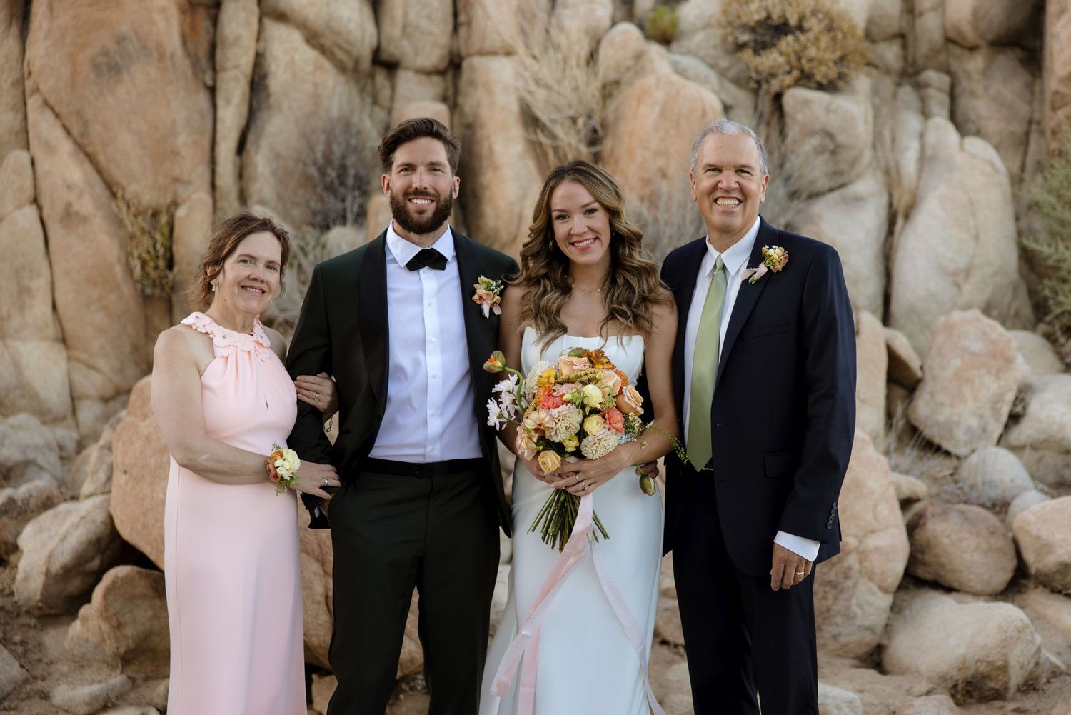 Wedding party in formal attire poses together in front of a rocky stone wall outdoors.