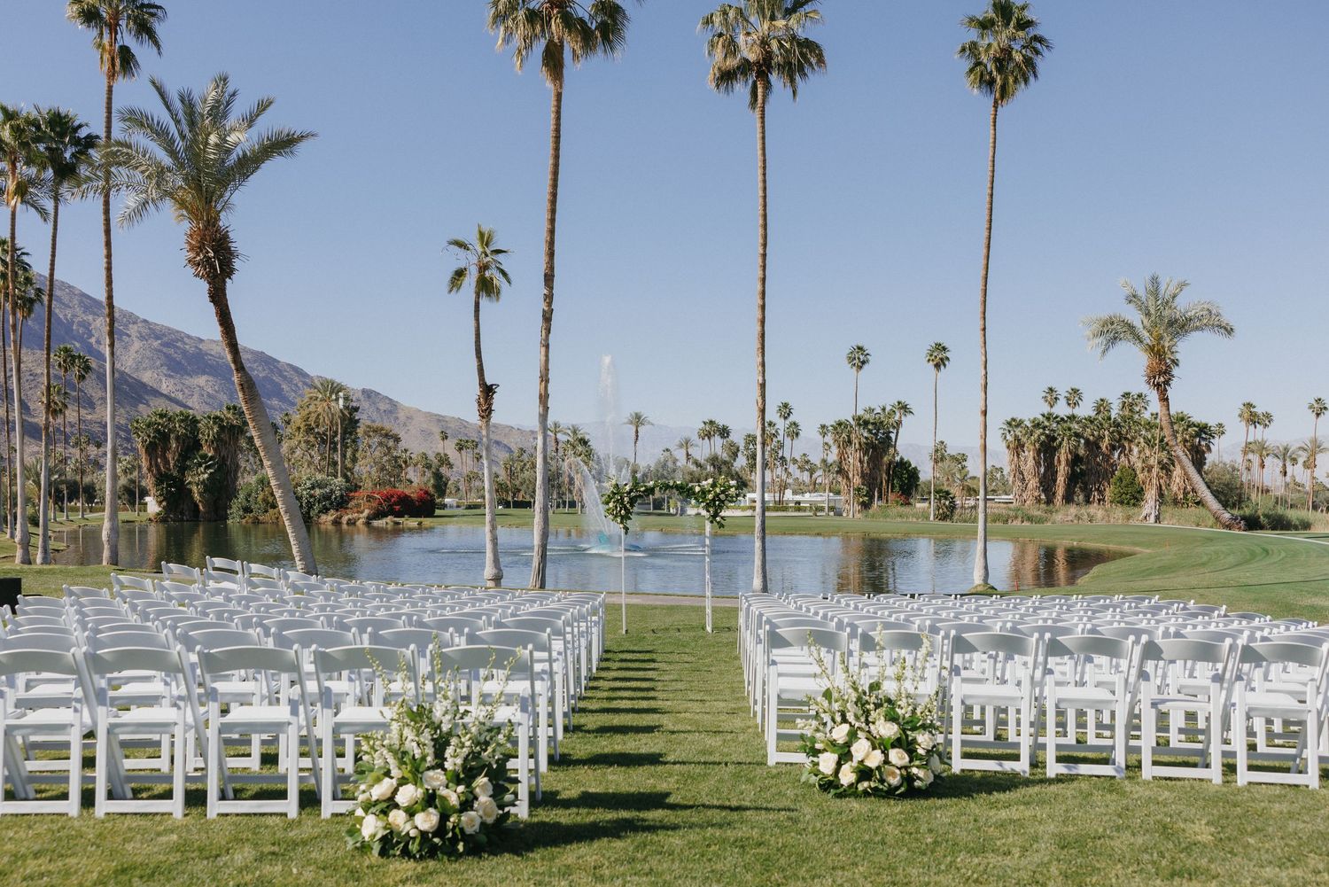 Outdoor wedding ceremony setup with white chairs, floral arrangements, palm trees, and mountain backdrop.