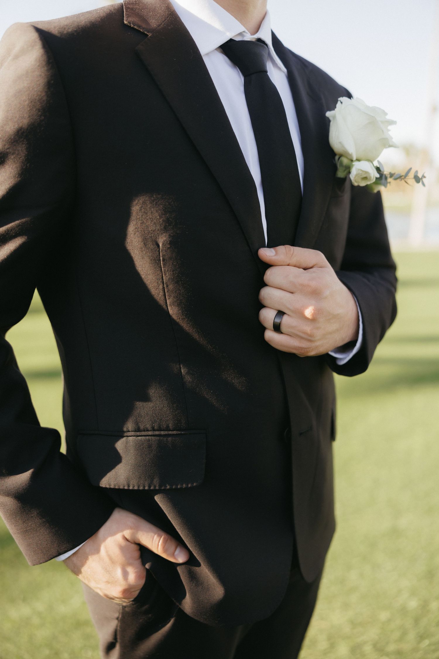 Groom in a black suit with white rose boutonniere and black tie adjusting his jacket on a sunny wedding day.