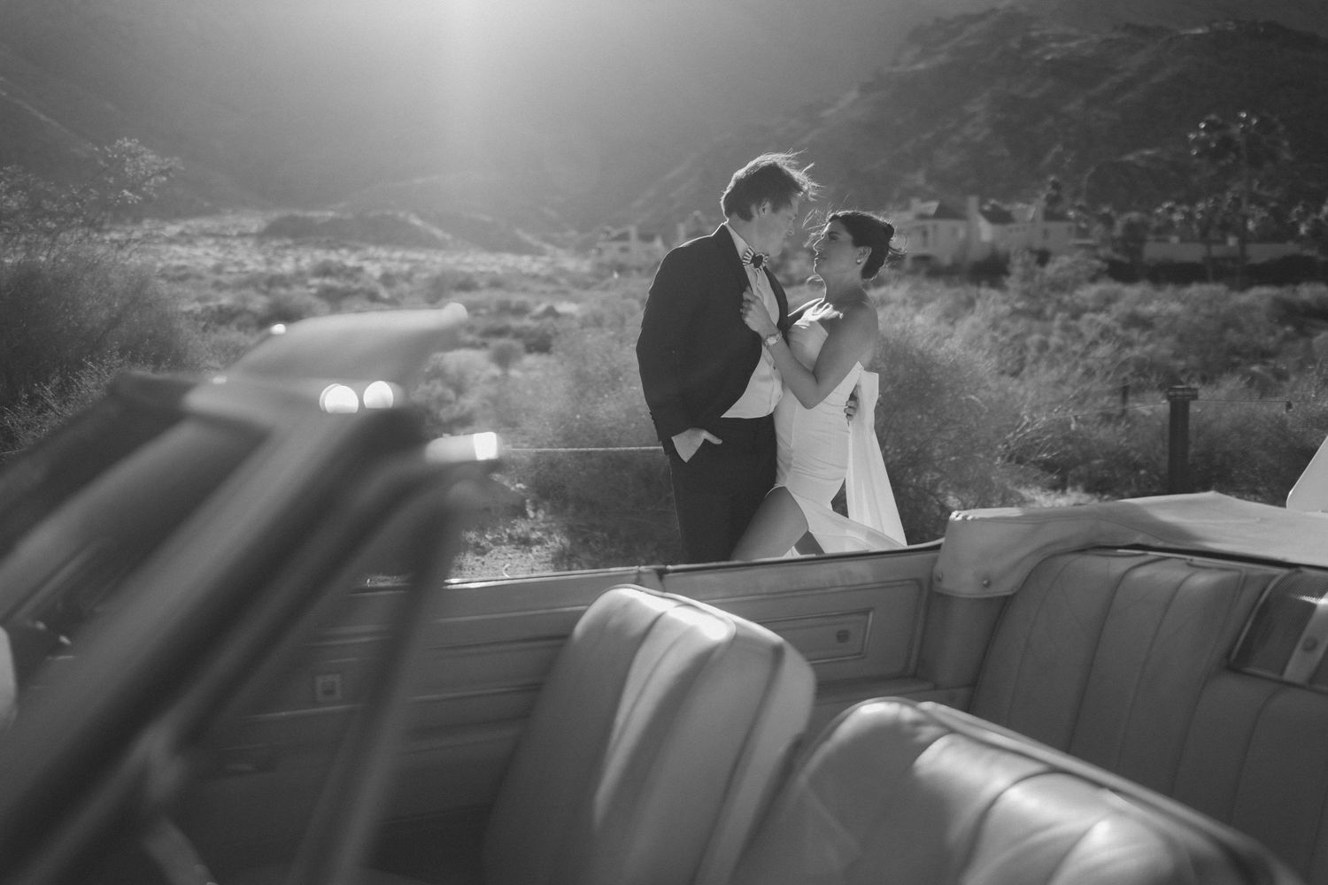 Couple sharing a romantic kiss in a vintage convertible car surrounded by desert mountains.