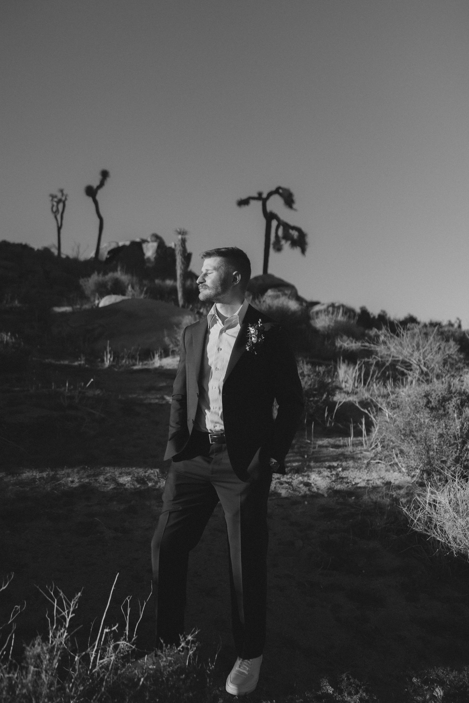 Black and white portrait of groom in dark suit with boutonniere standing in Joshua Tree desert landscape.