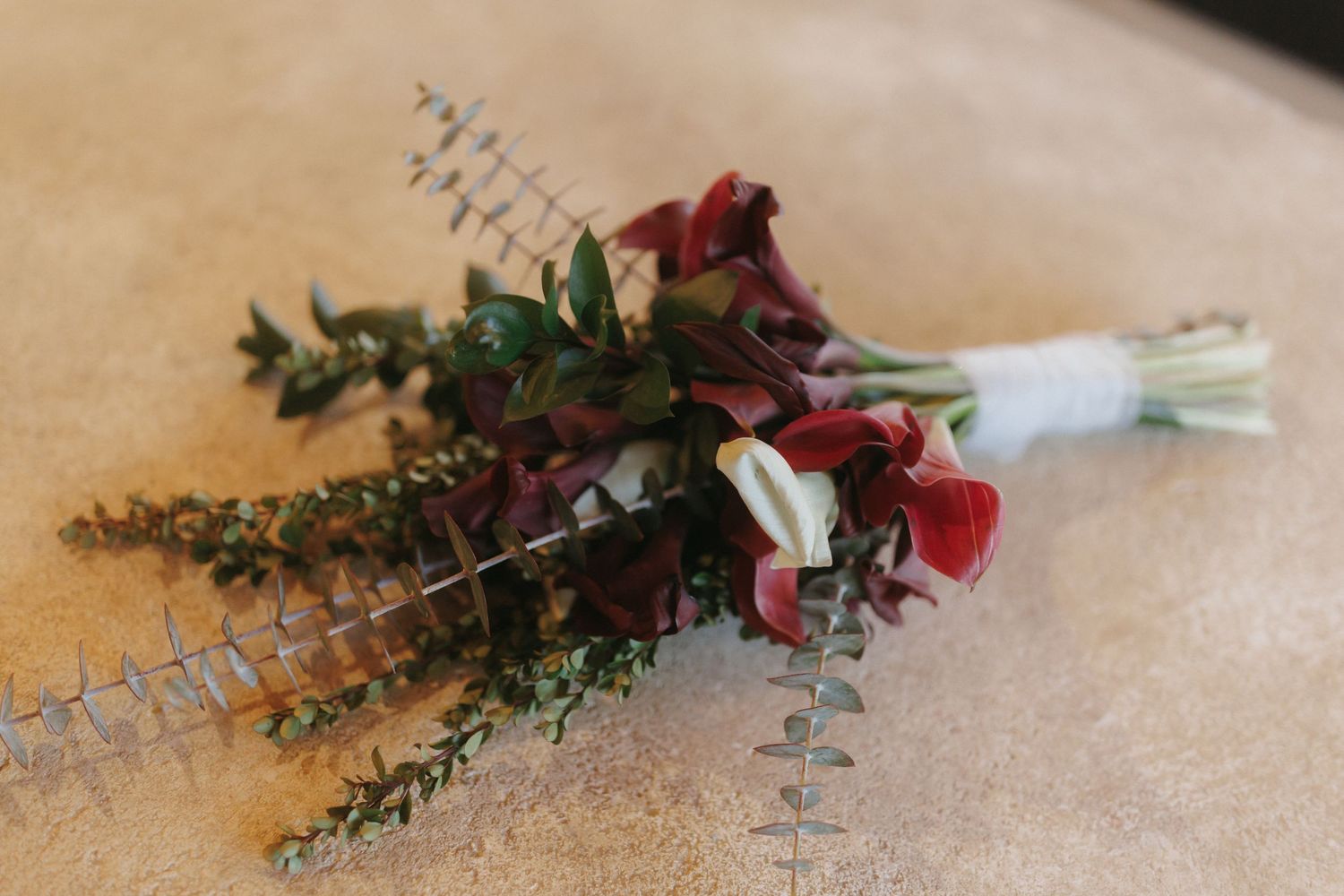 Elegant bridal bouquet with deep red flowers, greenery, and eucalyptus laid on a neutral surface.