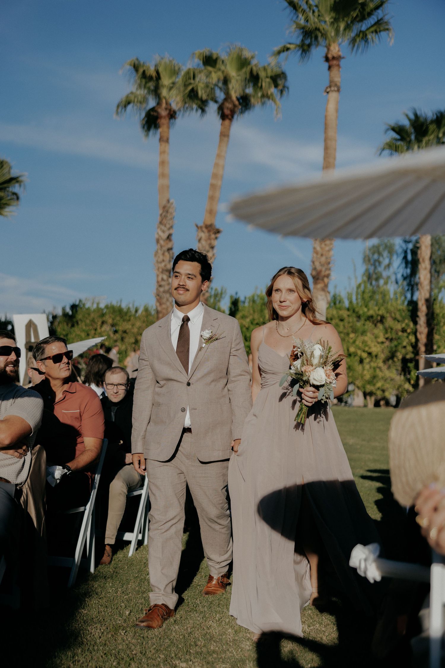 Wedding couple walks down outdoor aisle lined with palm trees at sunset garden ceremony.