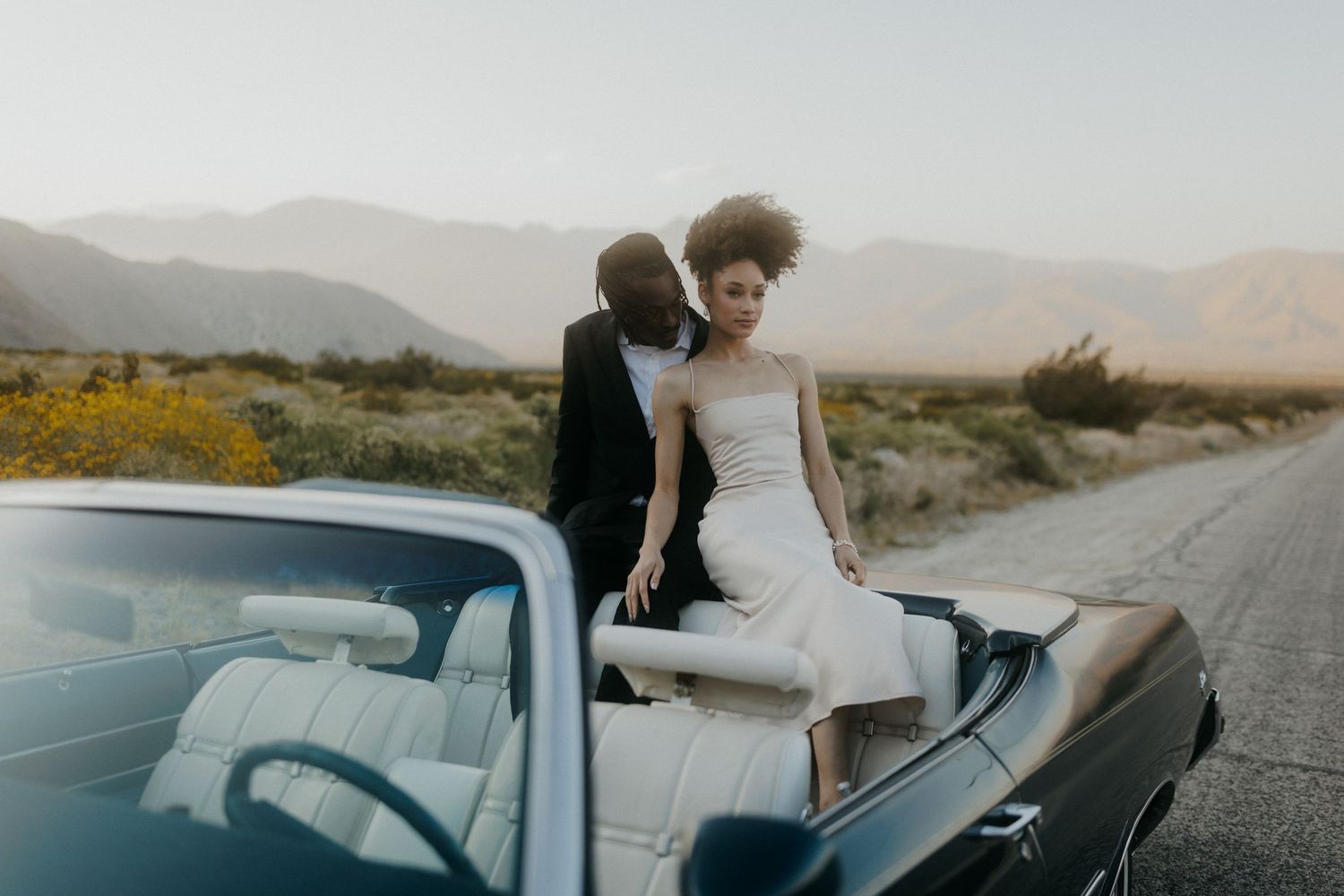 People celebrating in bridal attire across scenic desert landscapes with vintage car and mountains at sunset.