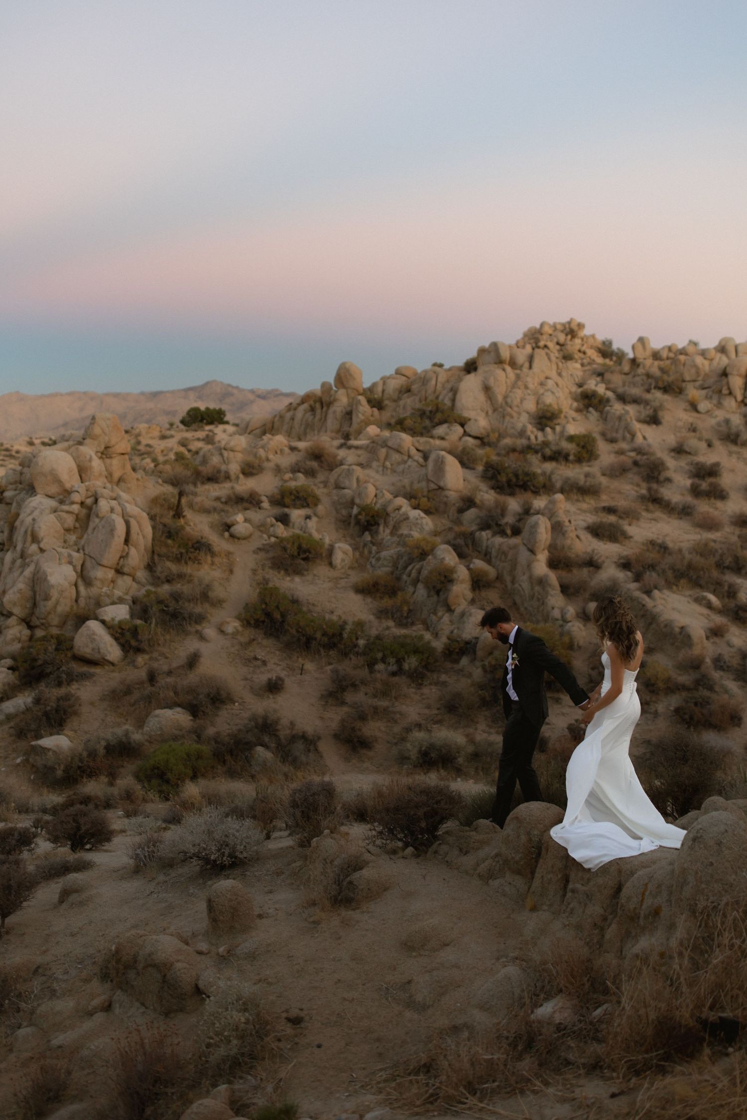 A desert landscape with rocky formations and silhouettes during sunset as a white dress billows in the wind.