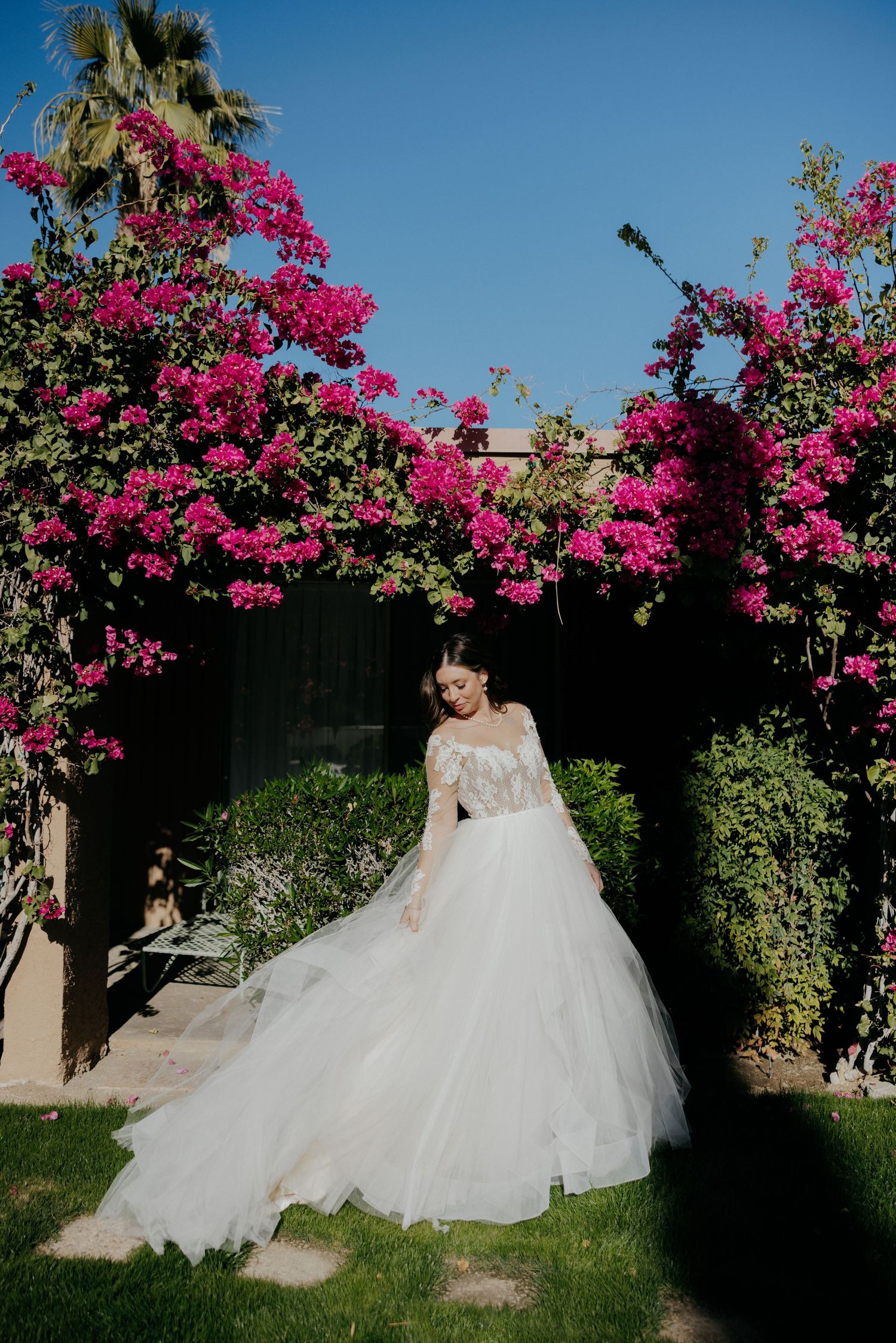 Beautiful white wedding gown under blooming pink bougainvillea arch in a garden with palm trees and blue sky.
