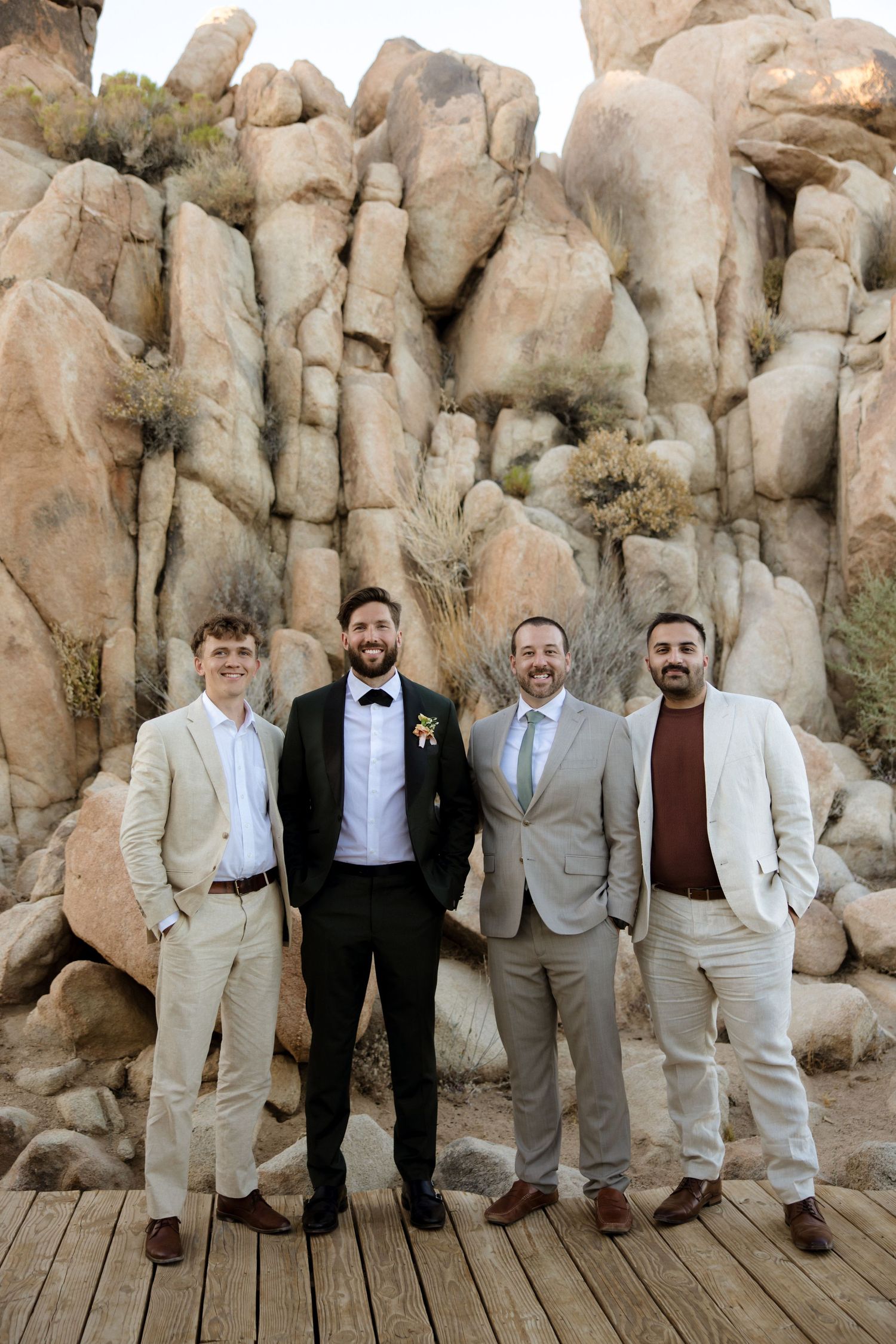 Groomsmen in tan suits and black bowties pose against desert rock formations at a scenic outdoor wedding venue.