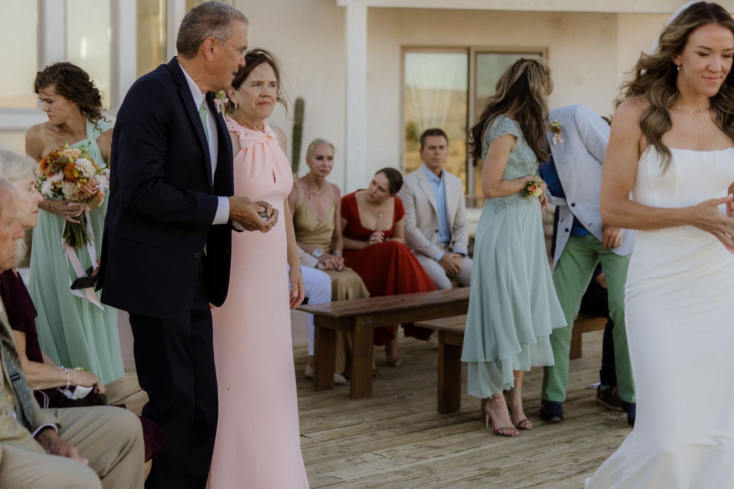 Wedding party gathers on a covered deck with attendees wearing pastel dresses in mint green and pink tones during ceremony.