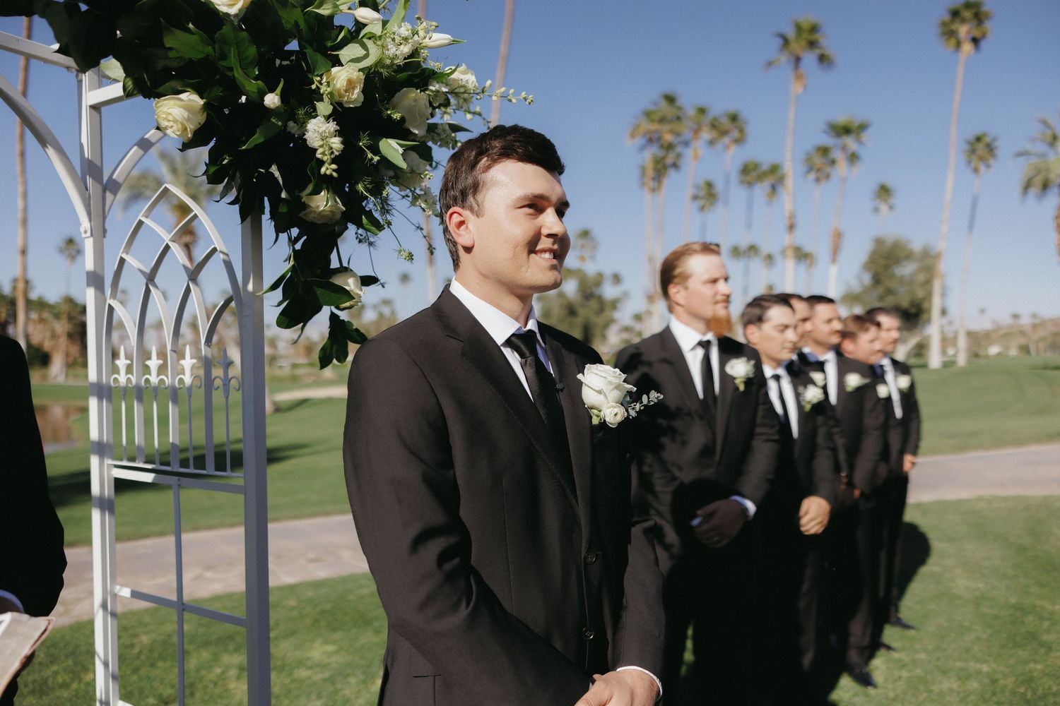 Groom in black suit smiles at altar with groomsmen lined up beside him at an outdoor Palm Springs wedding.