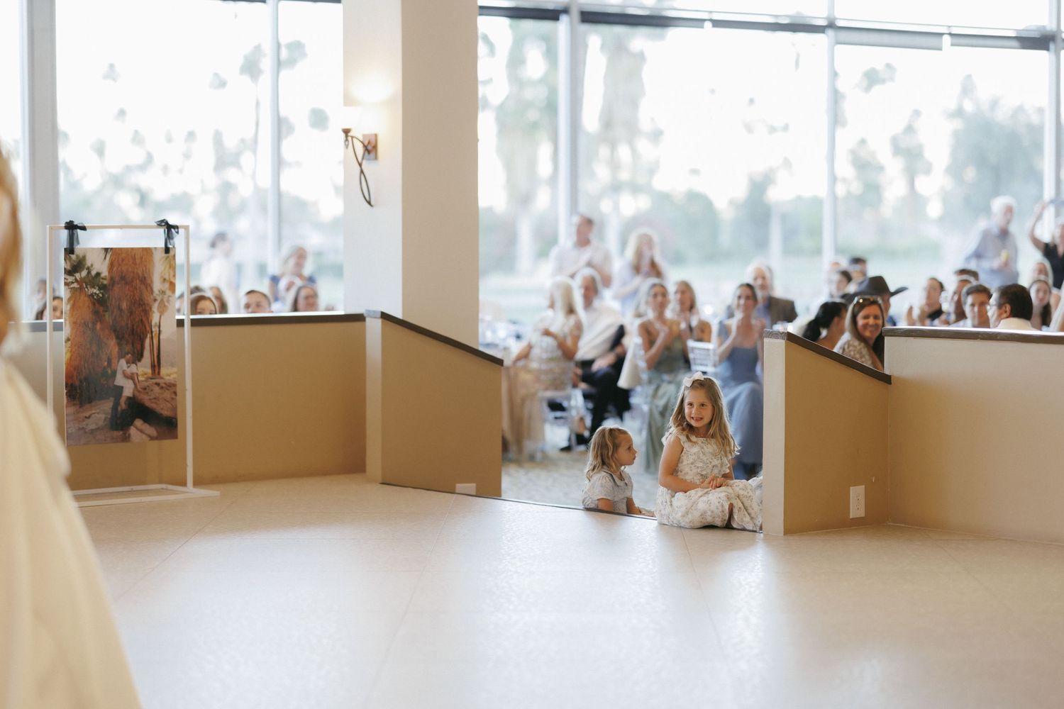 Wedding guests seated in bright venue watching ceremony, child sitting on floor in foreground.
