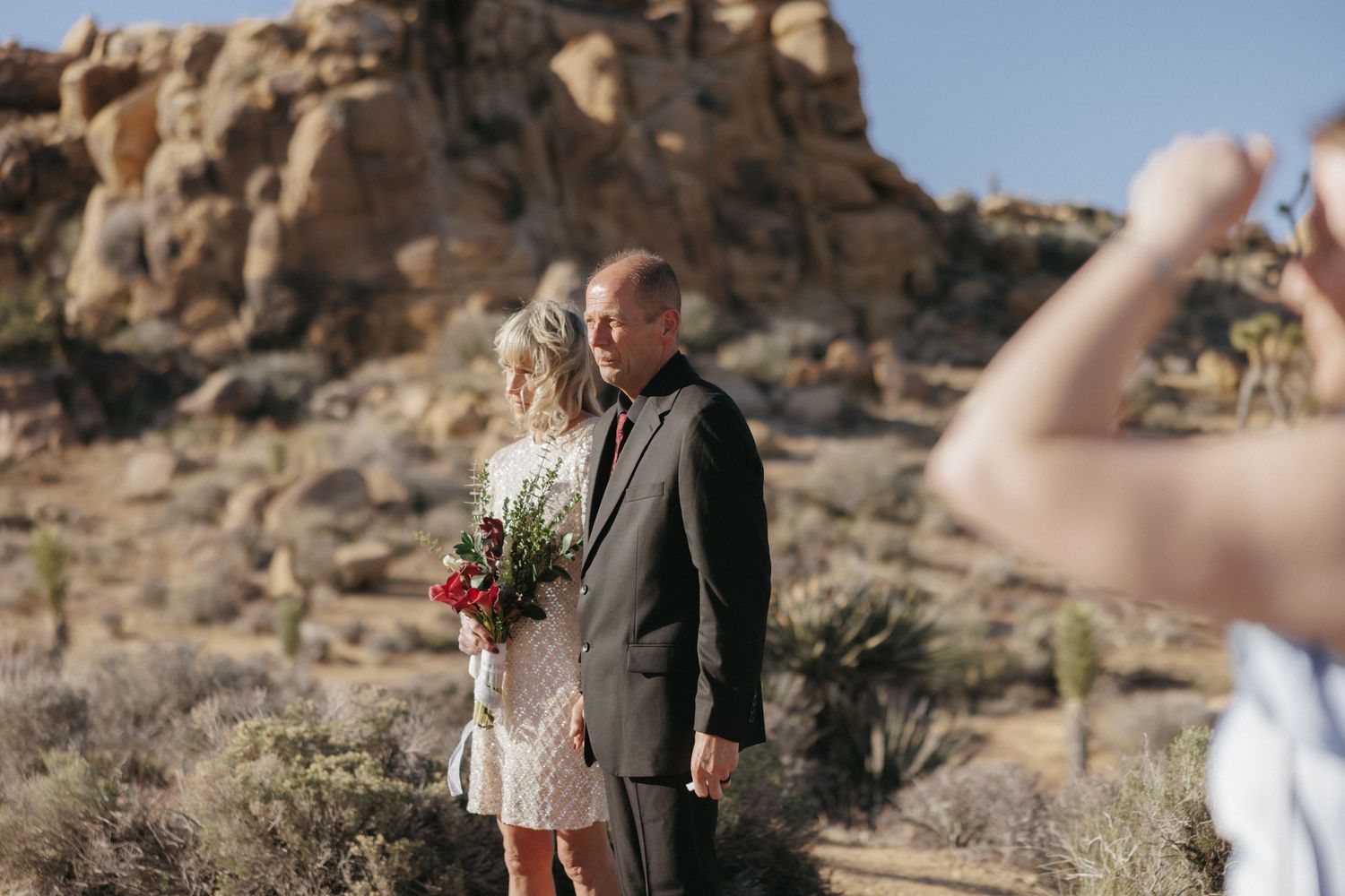 Bride and groom standing together at an outdoor desert wedding ceremony among Joshua Tree rock formations.