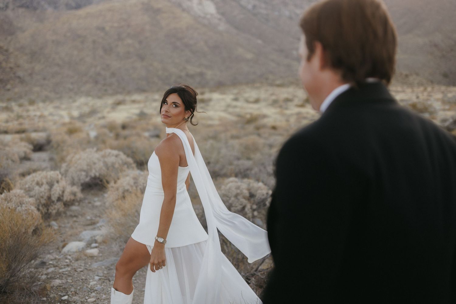 Bride in backless white gown glances back at groom while walking through a rocky desert mountain landscape.