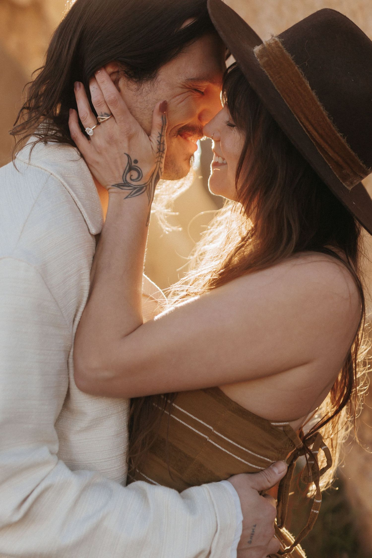 Romantic couple sharing an intimate moment at golden hour, woman in brown hat with tattoo embracing man in warm sunlight.