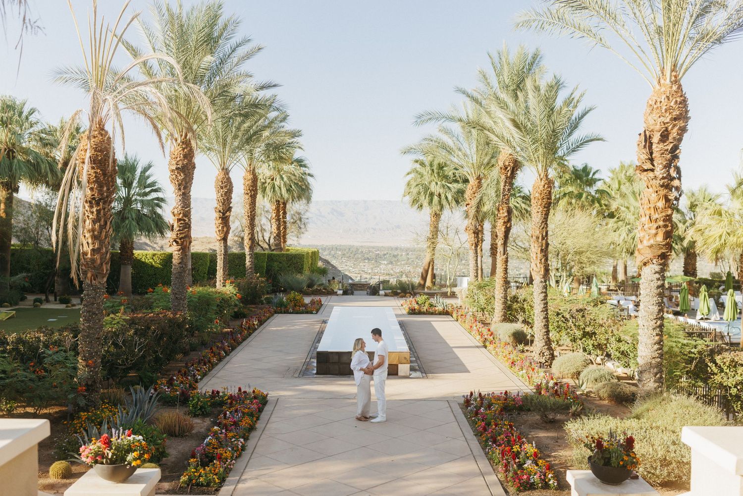 Couple embraces on elegant palm-lined pathway at desert resort with mountain views and colorful garden beds.