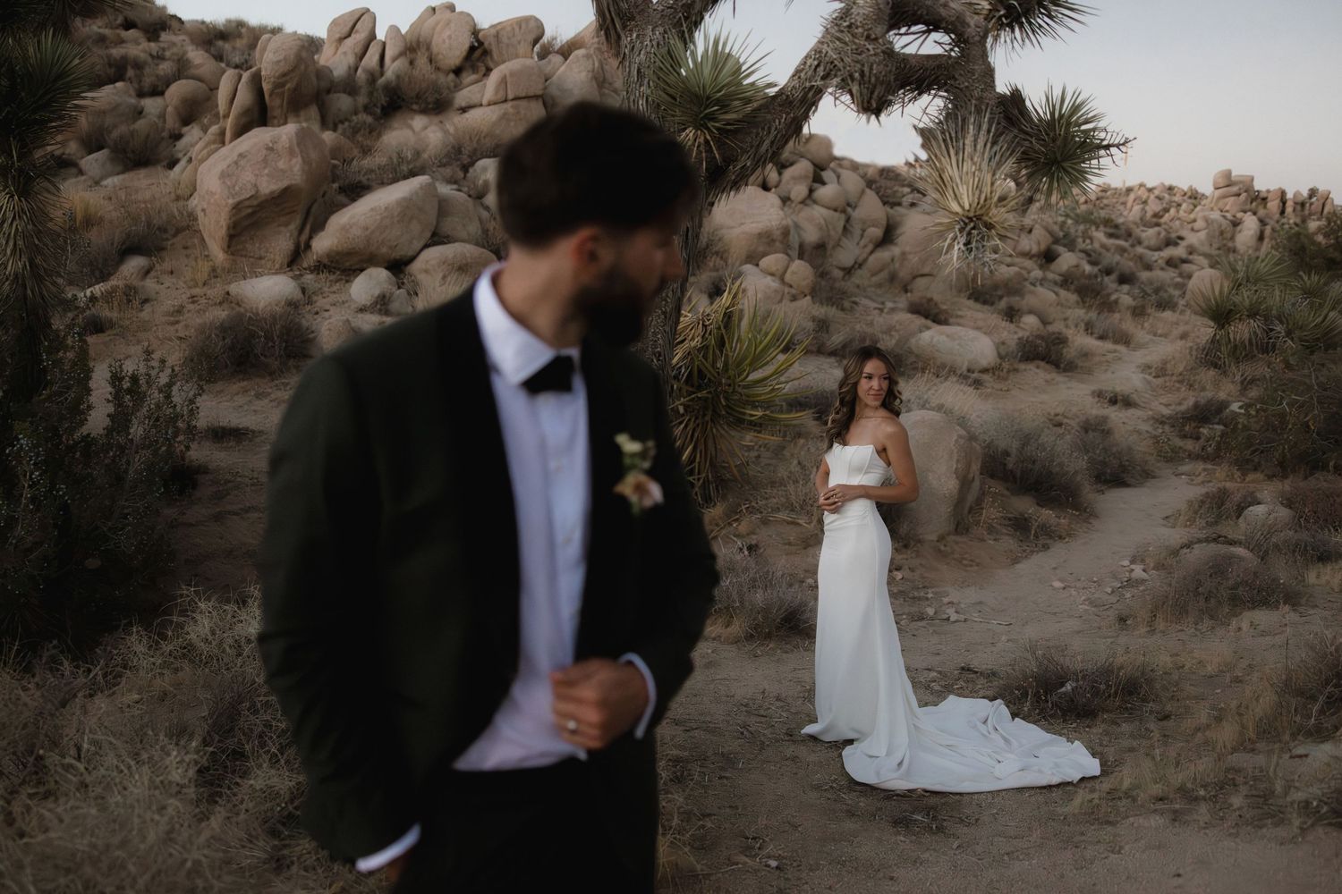 A bride in a white gown and groom in a dark suit pose among desert boulders and Joshua trees at their outdoor wedding.