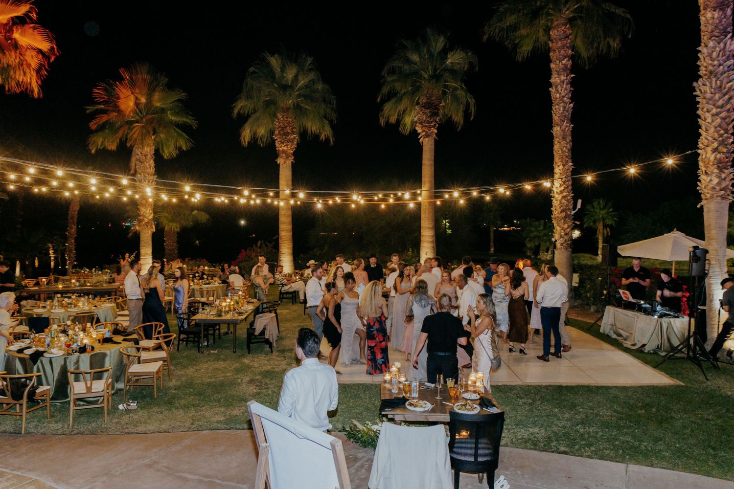 Wedding guests enjoying an outdoor evening reception under string lights and palm trees at a tropical venue.