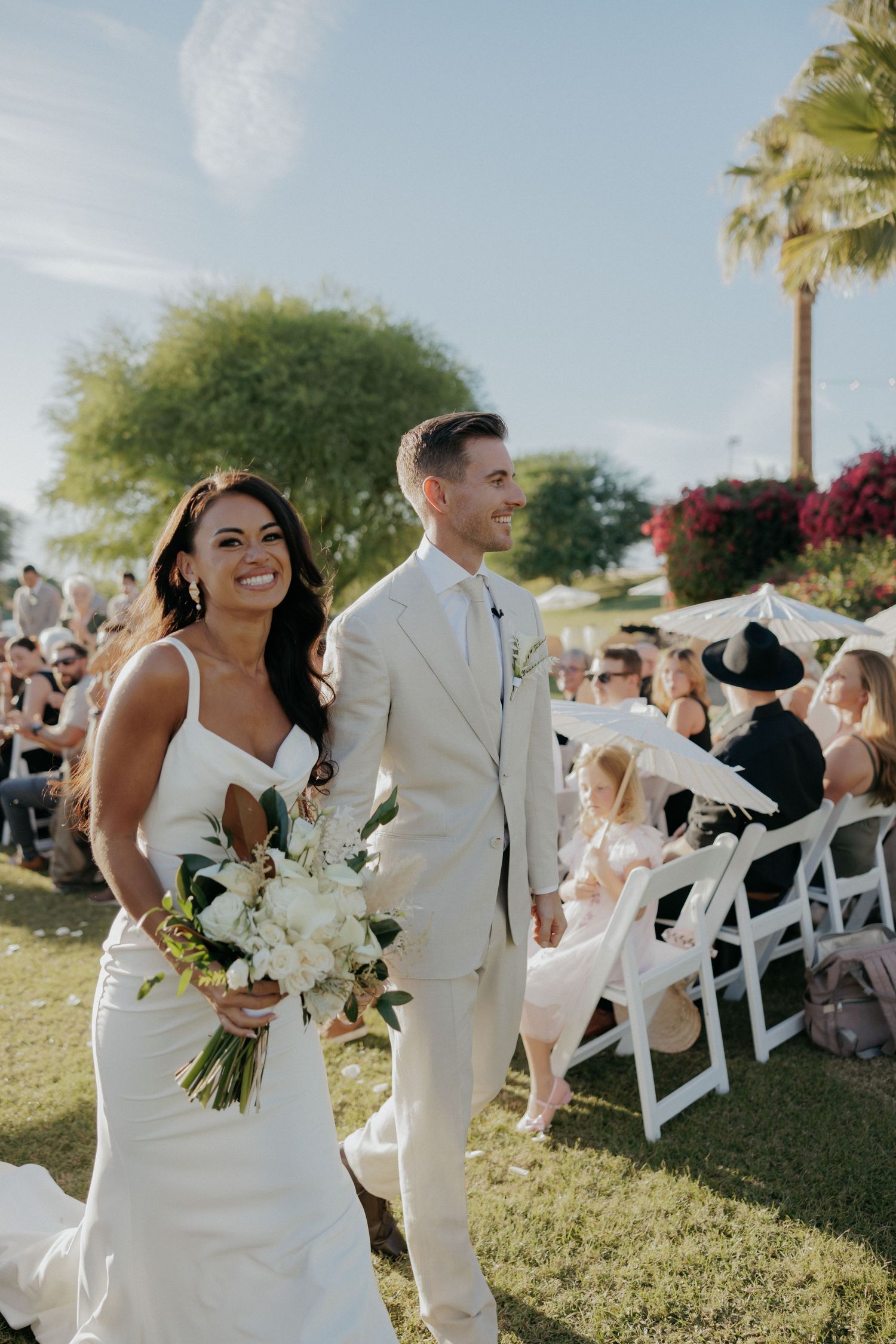 A wedding ceremony held outdoors with white chairs and palm trees in a scenic garden setting.