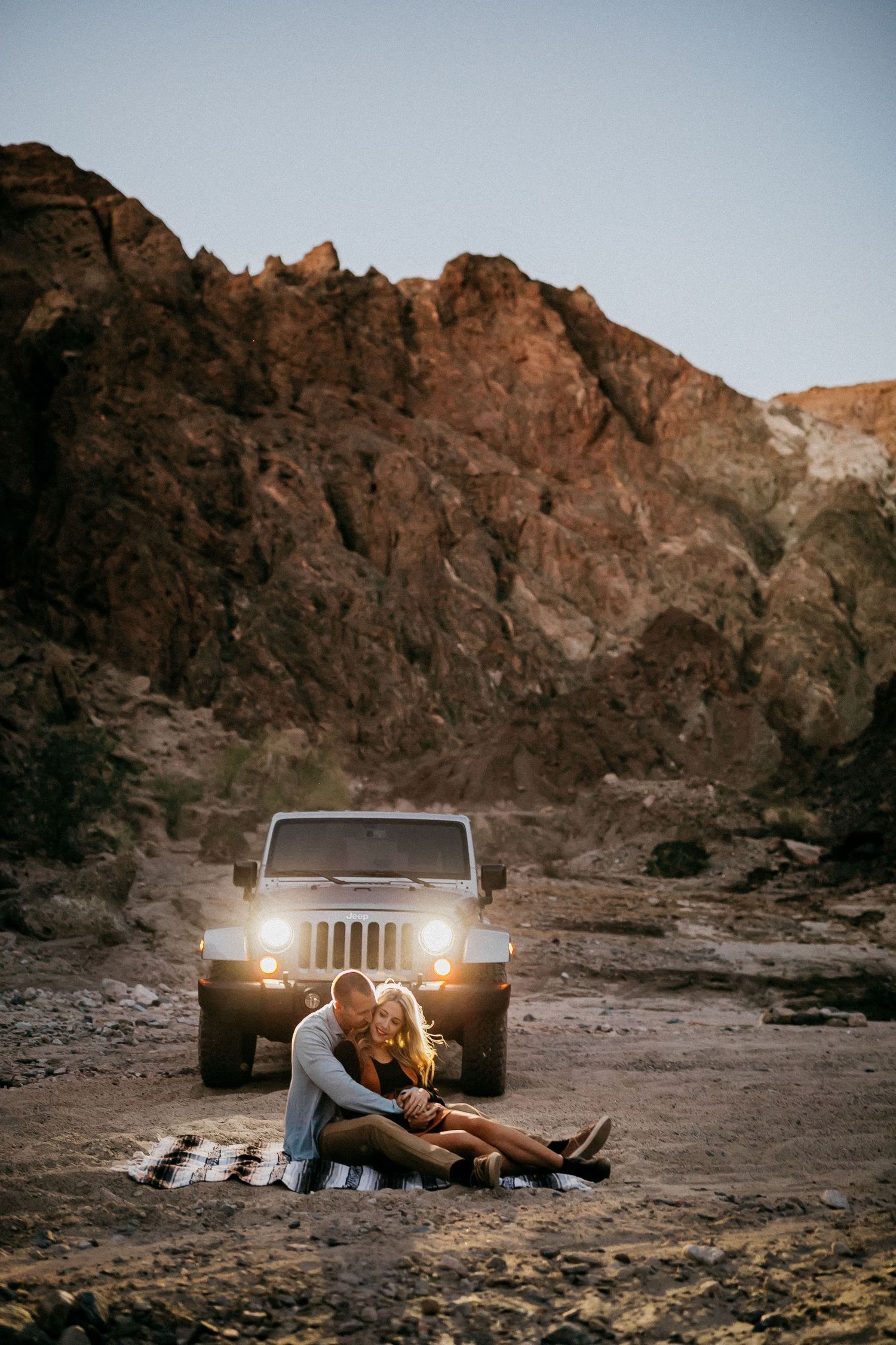 Couple enjoying a campfire next to parked Jeep with headlights on in rugged desert landscape during sunset.