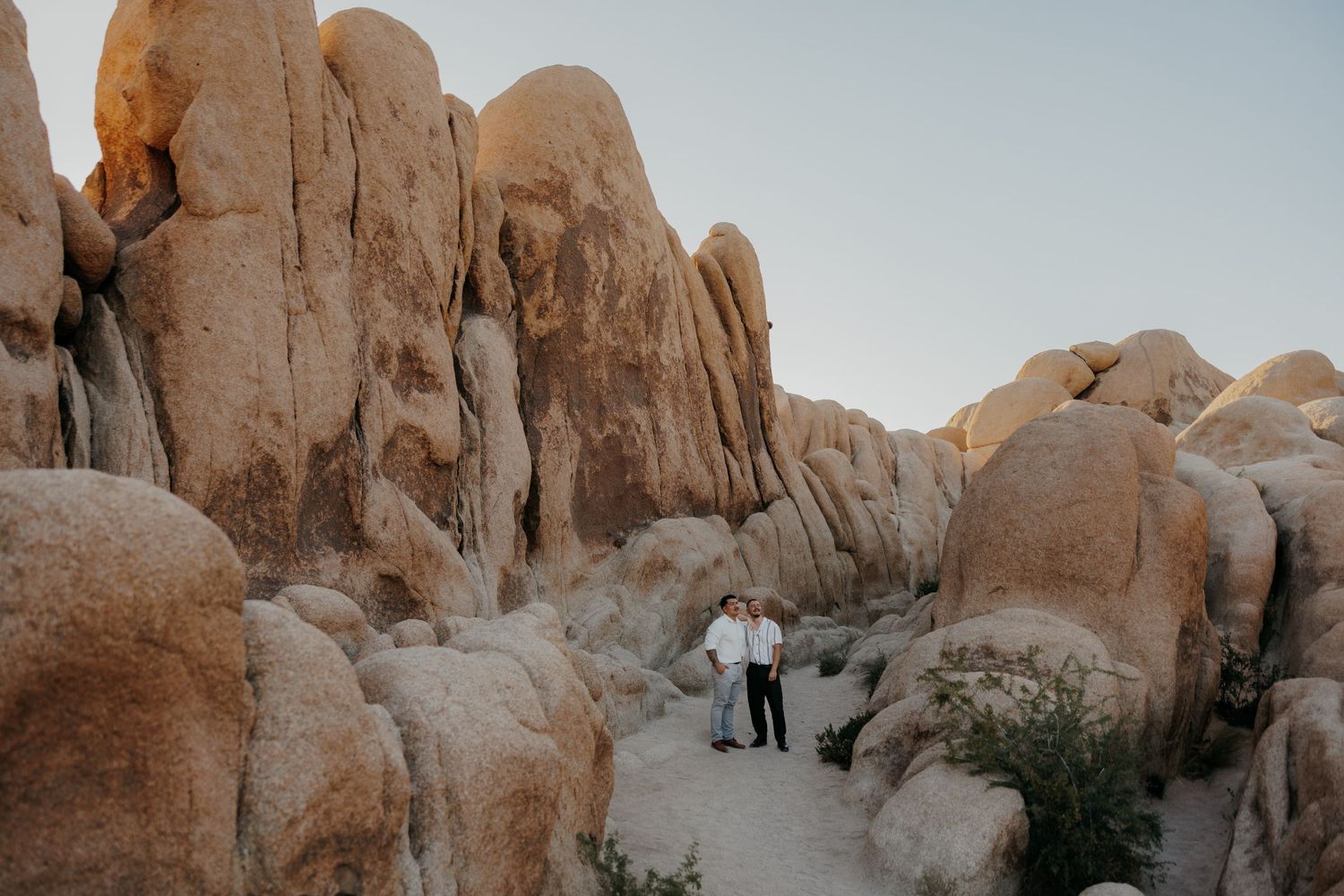 Two people walking between towering rock formations at sunset in Joshua Tree National Park.