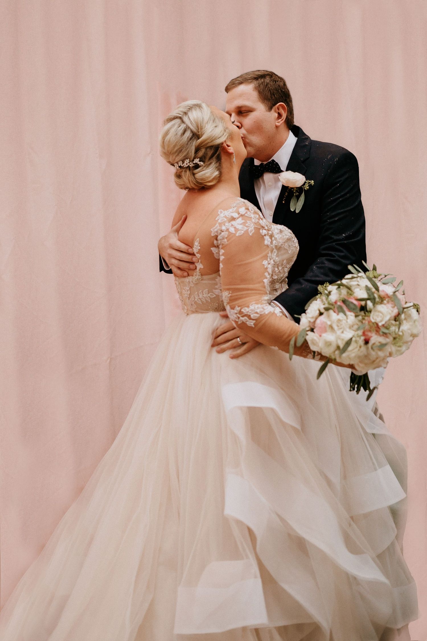 Romantic wedding day moment of bride and groom embracing in formal attire against blush pink backdrop.