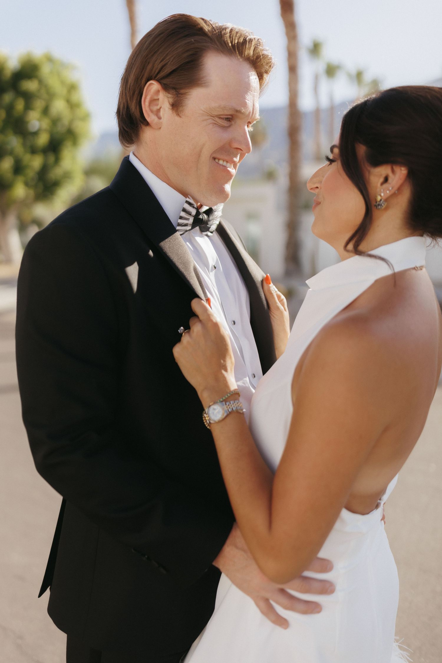 Bride and groom share a joyful moment, groom in black tuxedo with bow tie, bride in white halter gown, palm trees behind.