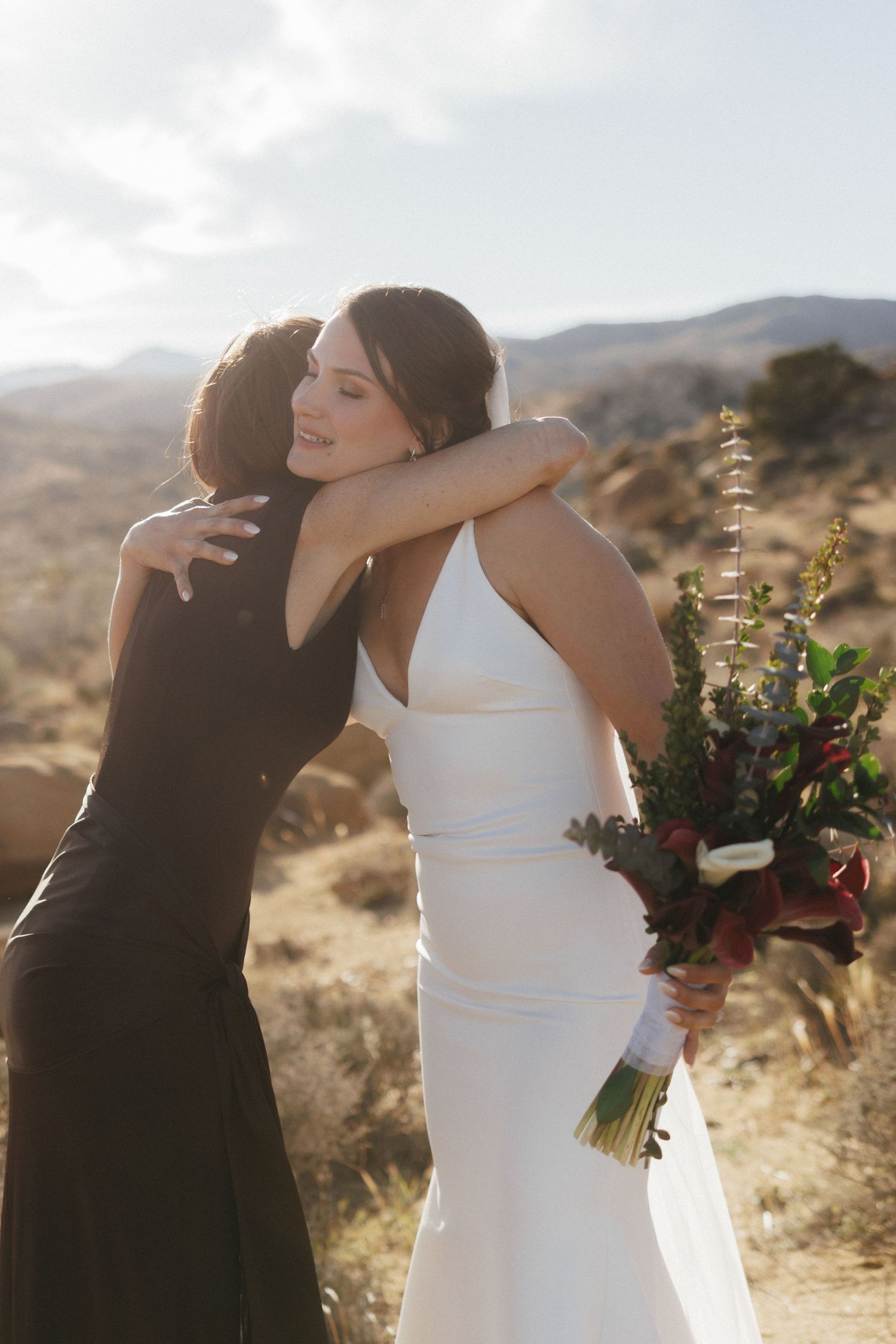 Bride in white gown hugs guest while holding bouquet in desert setting.