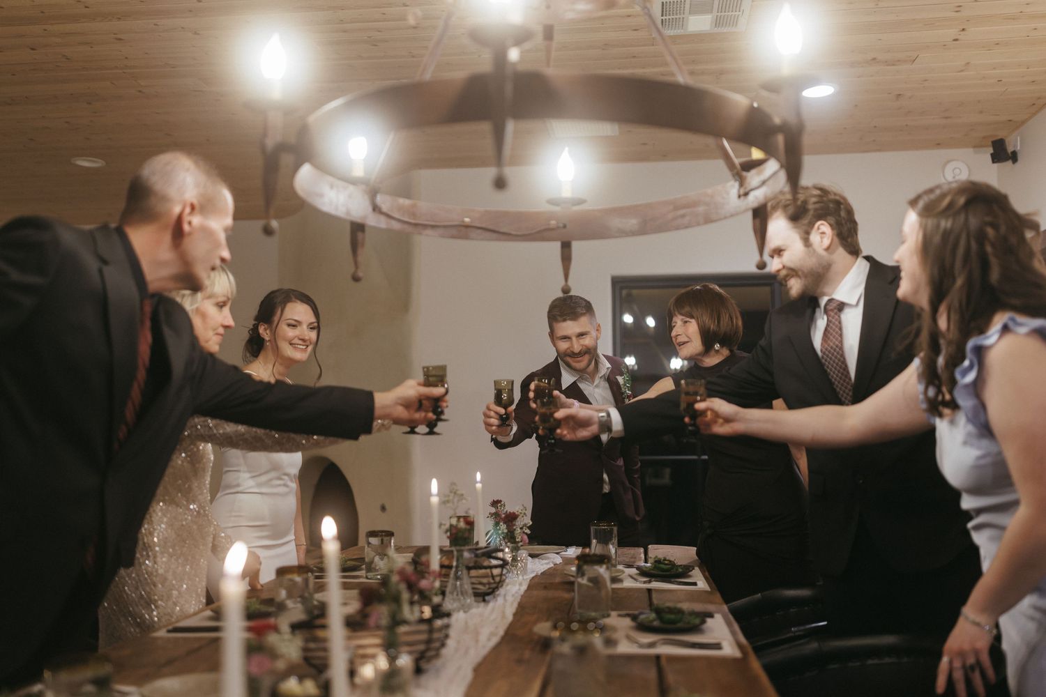 Wedding guests raise glasses in a warm candlelit toast around a rustic decorated reception table indoors.