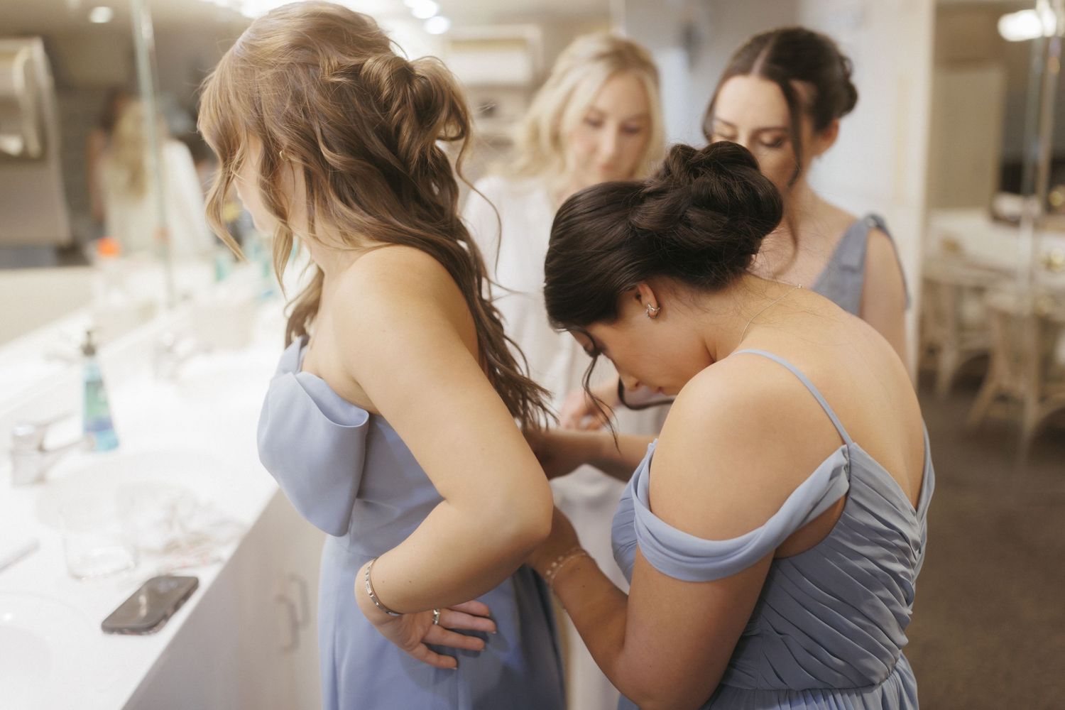 Bridesmaids in blue dresses helping each other get ready before a wedding ceremony indoors.