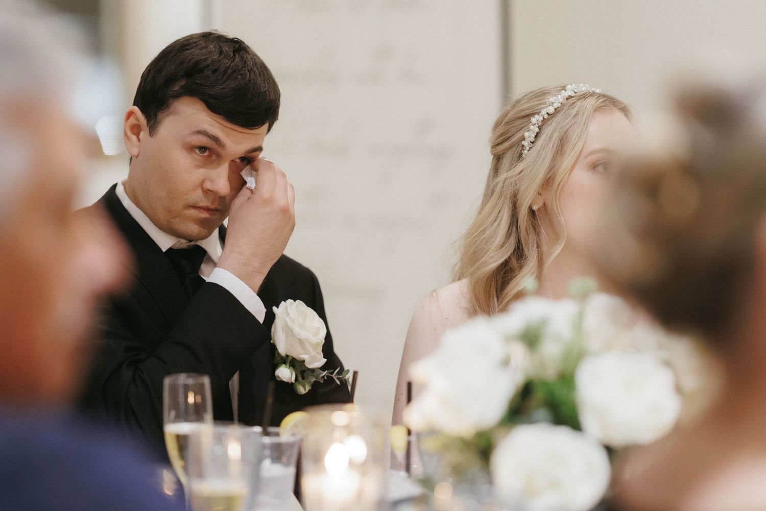 Emotional groom wipes a tear at wedding reception table beside bride with white floral centerpieces and candles.