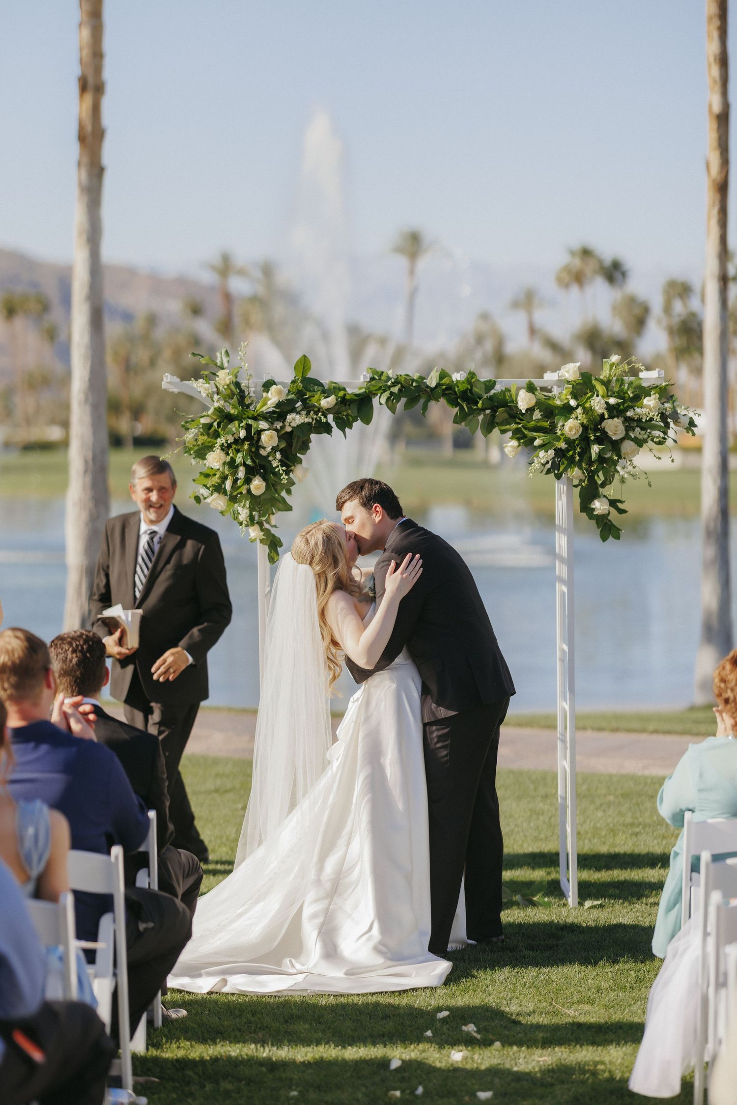 Bride and groom share first kiss under floral arch at outdoor wedding ceremony by a lake with fountain.