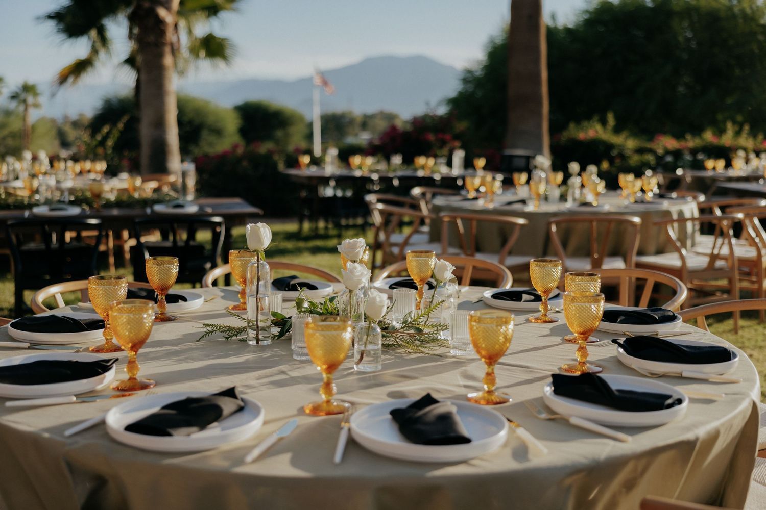 Elegant outdoor wedding reception tables with white linens and place settings arranged under palm trees at sunset.