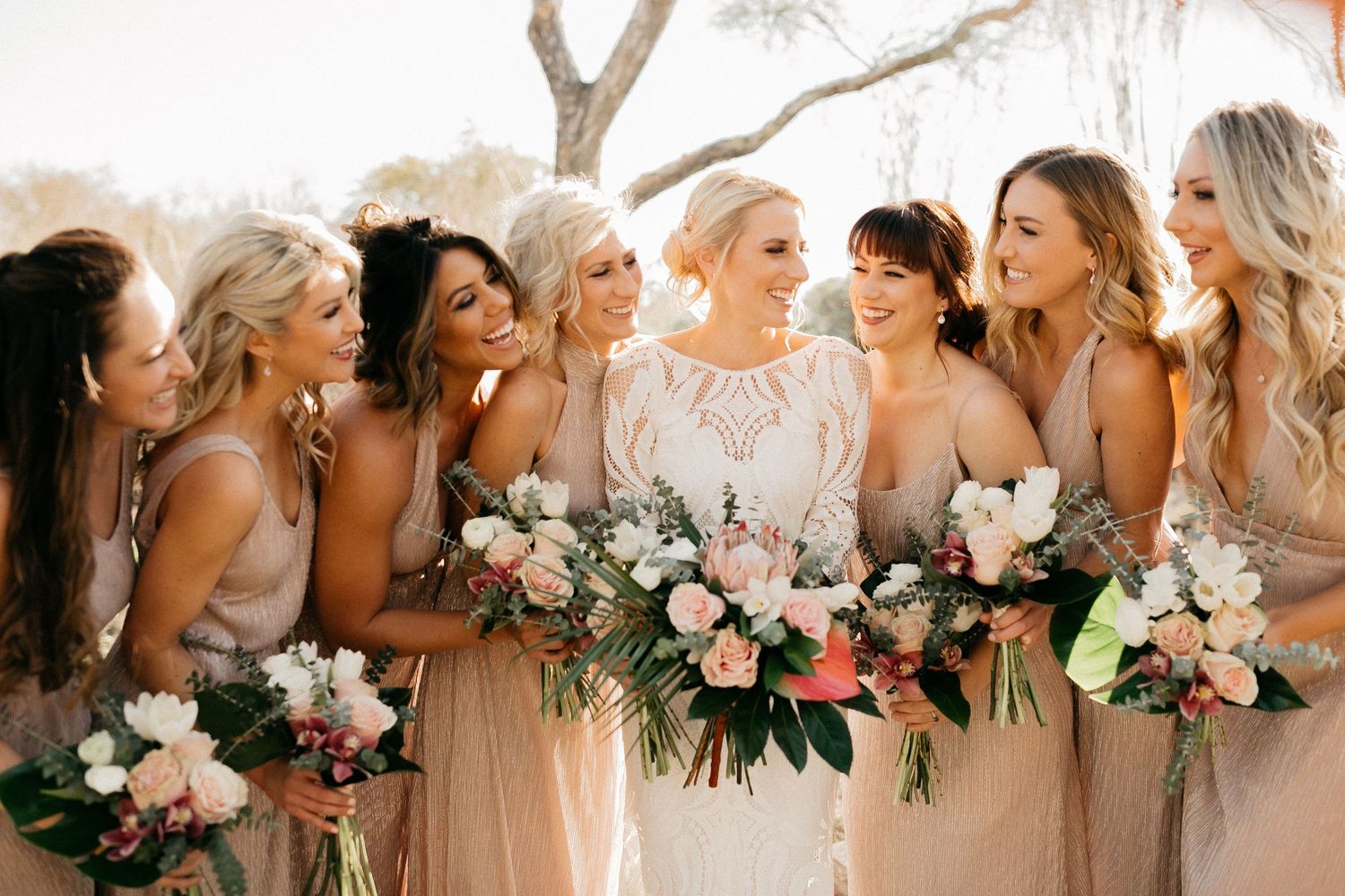 Bridesmaids in blush pink dresses holding floral bouquets share laughter and smiles in a candid outdoor wedding photo.