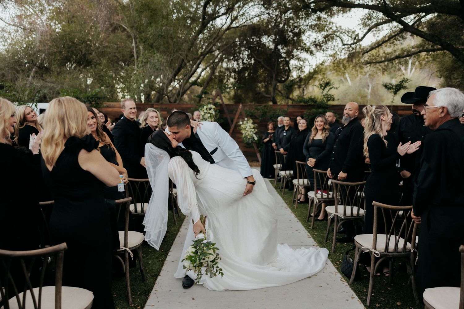 Wedding guests dance and celebrate during an outdoor reception under trees and string lights.