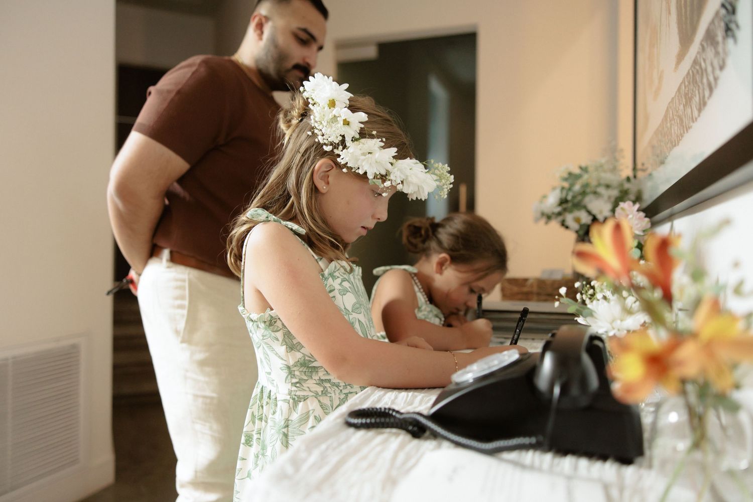 Wedding party preparing together in a sunlit room with floral arrangements and elegant dresses.