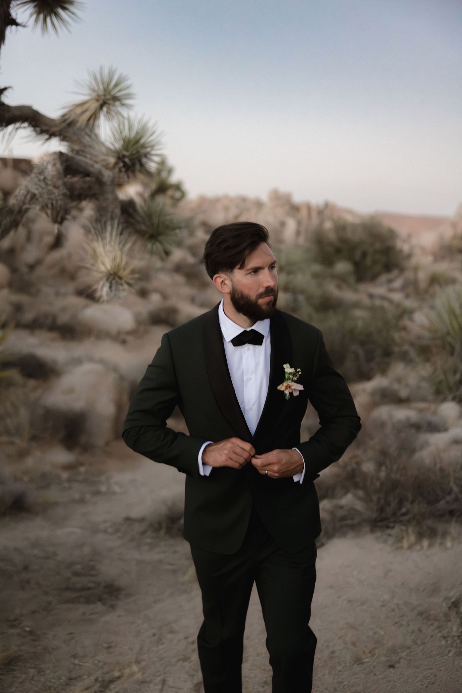A groom in a black tuxedo poses thoughtfully in a desert landscape with yucca plants at sunset.