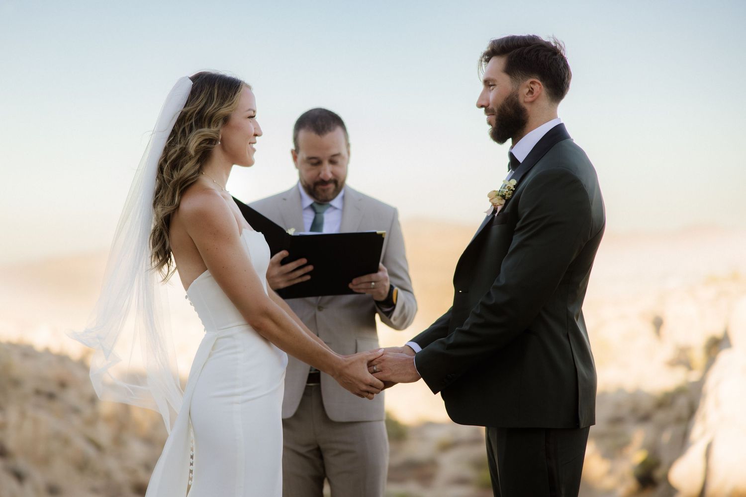 Outdoor wedding ceremony takes place on desert cliff during golden sunset hour with couple exchanging vows.