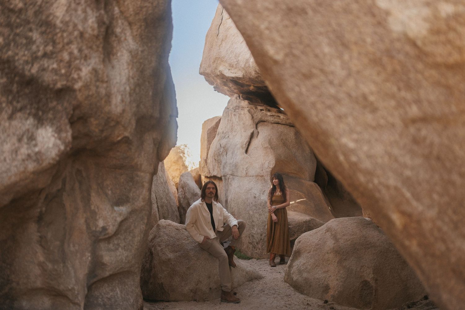 Couple posing among large boulders in a desert canyon, woman in flowing dress, man seated, warm golden light.