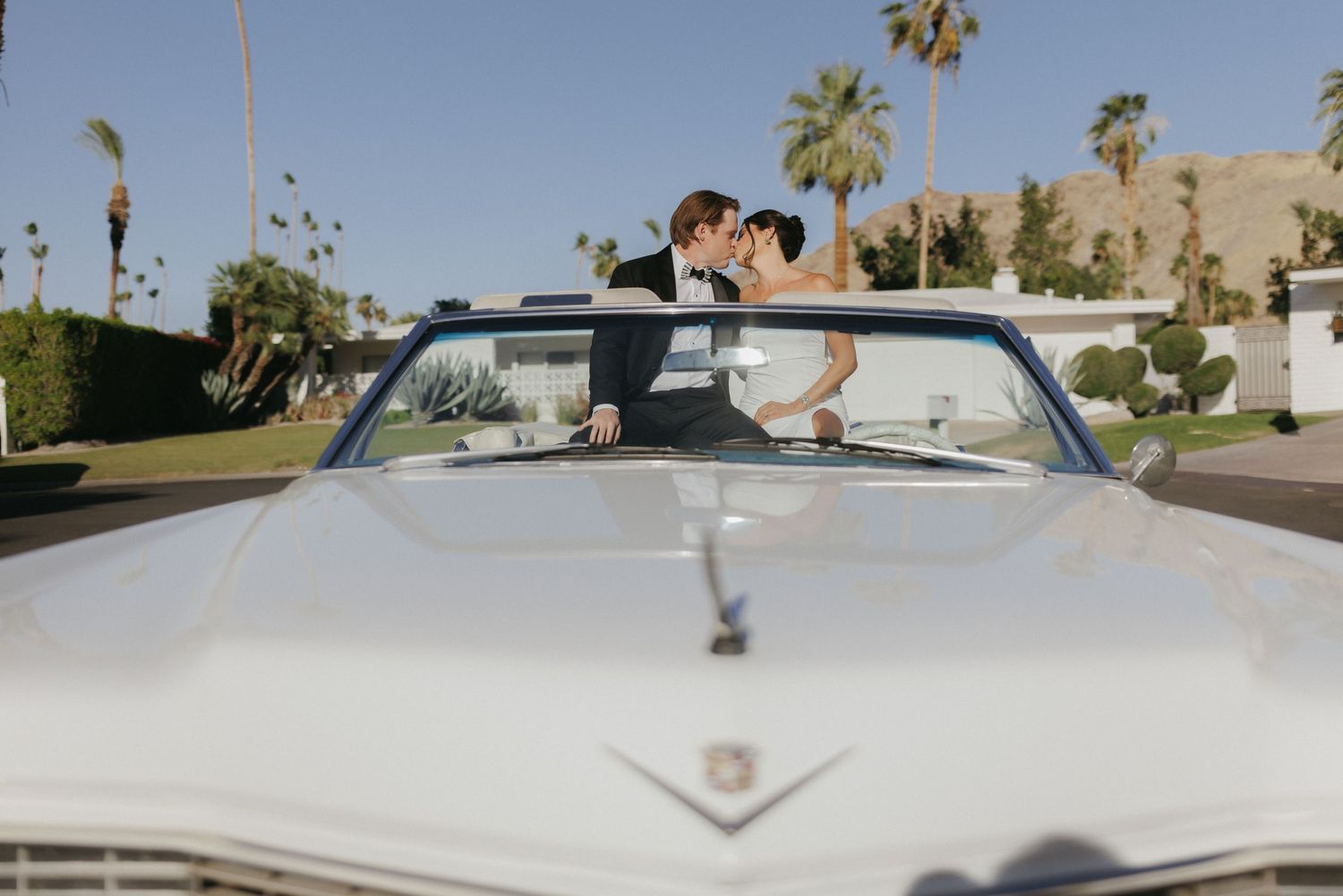 Bride and groom kissing behind a classic white Cadillac convertible with palm trees in background.