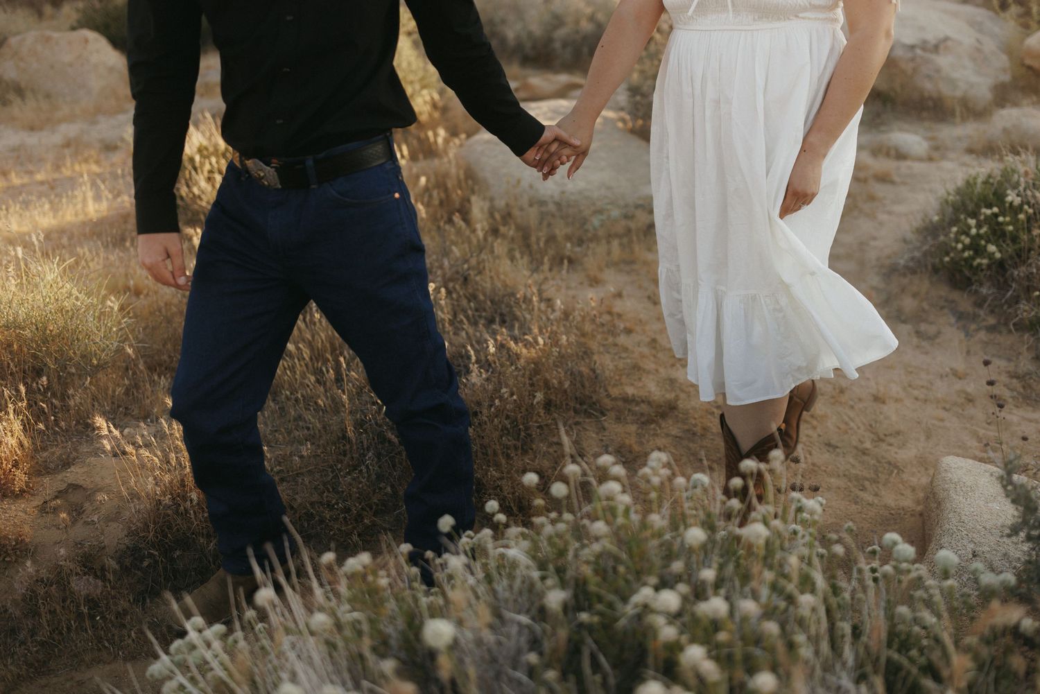 Couple holding hands in a desert field, man in dark jeans and woman in flowing white dress among wildflowers.