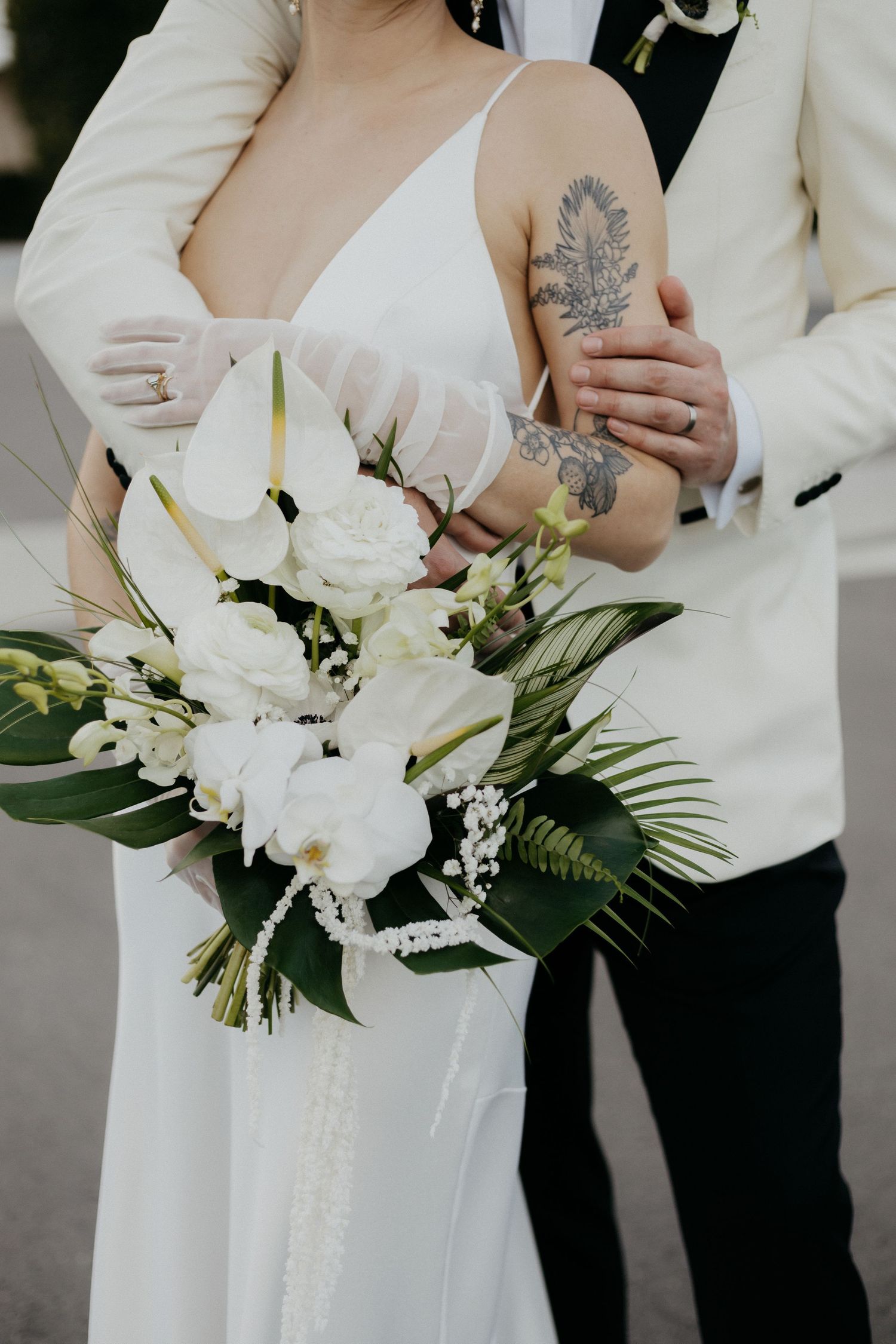 A bride in a white dress holds an elegant white floral bouquet with palm leaves while showing an ornate arm tattoo.