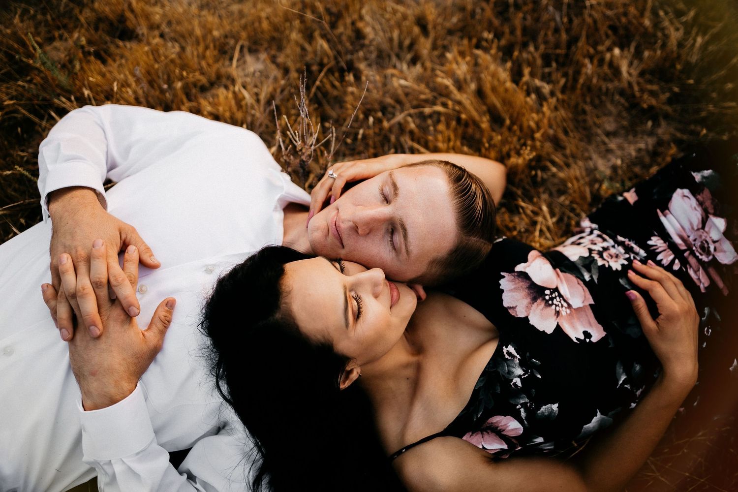 Couple lying together on autumn ground with fall foliage, one wearing a floral dress and other in white shirt.