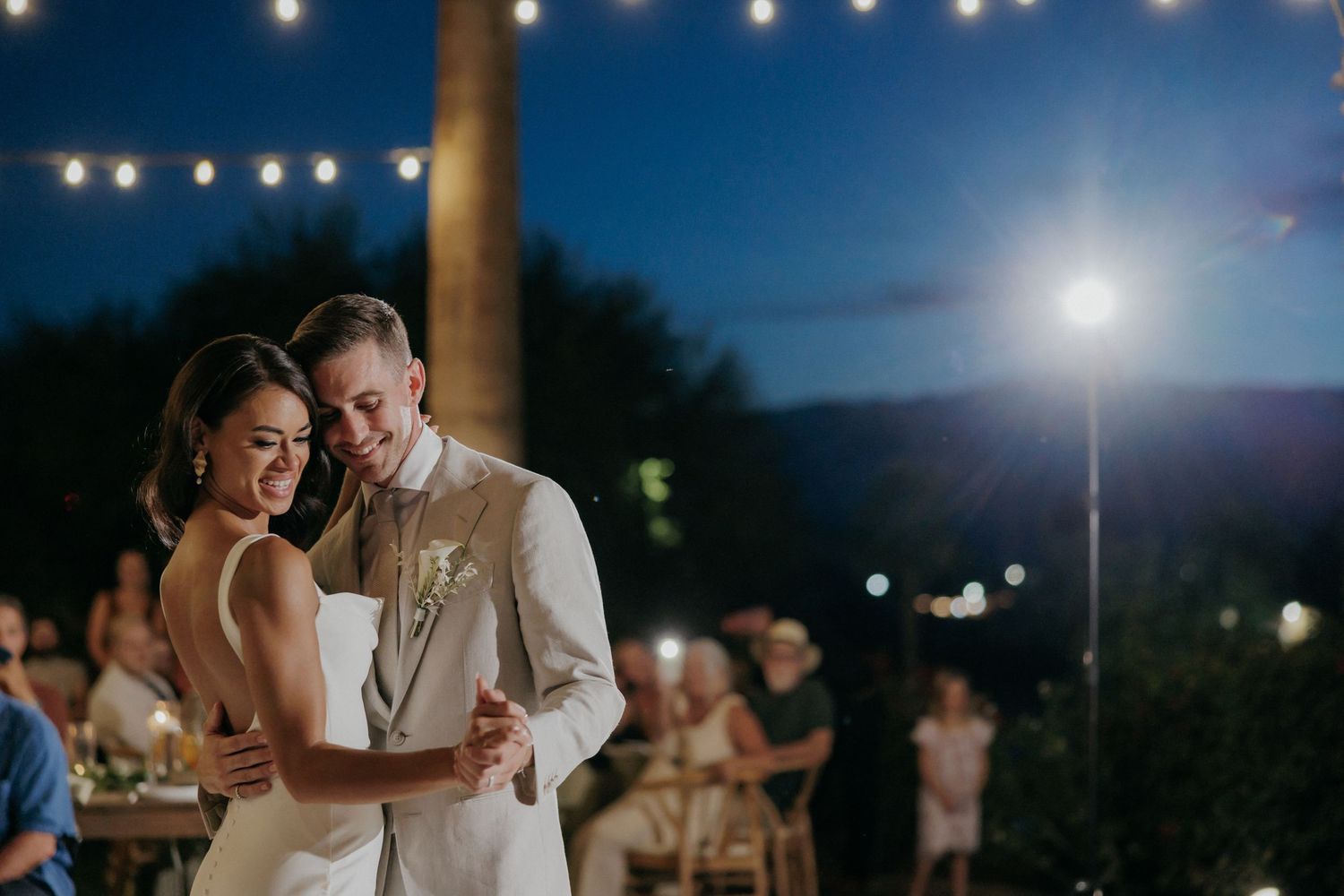 Romantic newlywed couple dances together under twinkling lights at an outdoor evening reception.