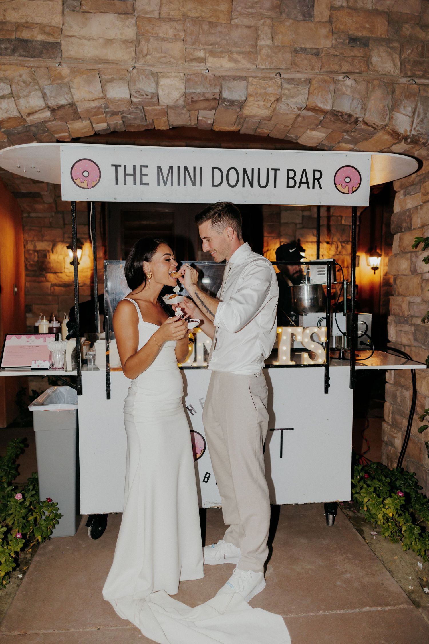 A couple dances in front of a rustic stone wall next to a mini donut bar at a wedding reception.