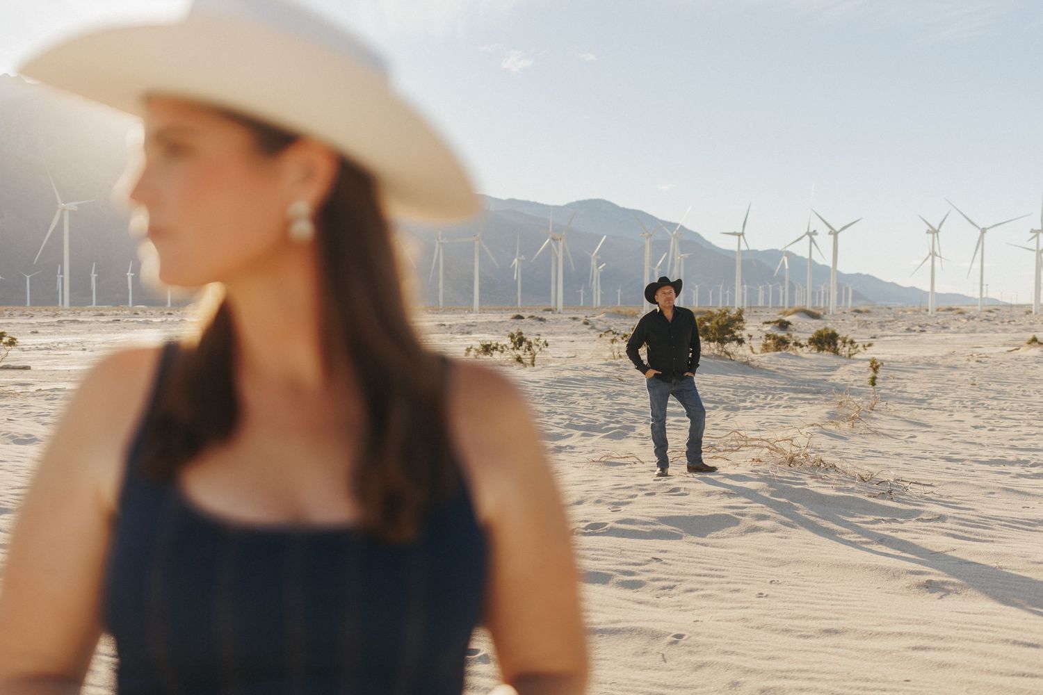 Couple in cowboy hats poses in desert with wind turbines and mountains in background.