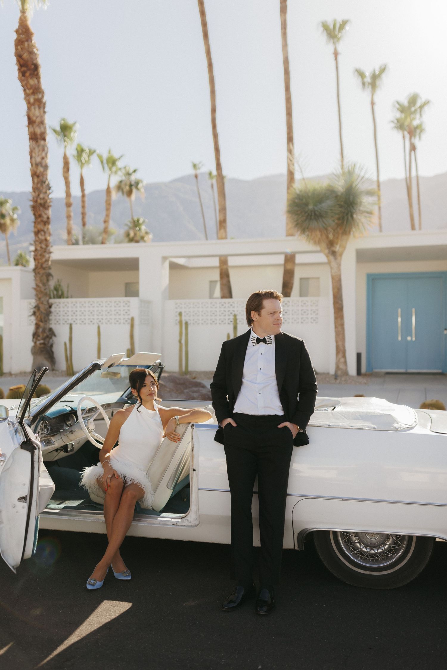 Bride and groom posing by a white vintage convertible in Palm Springs, with palm trees and mountains in the background.